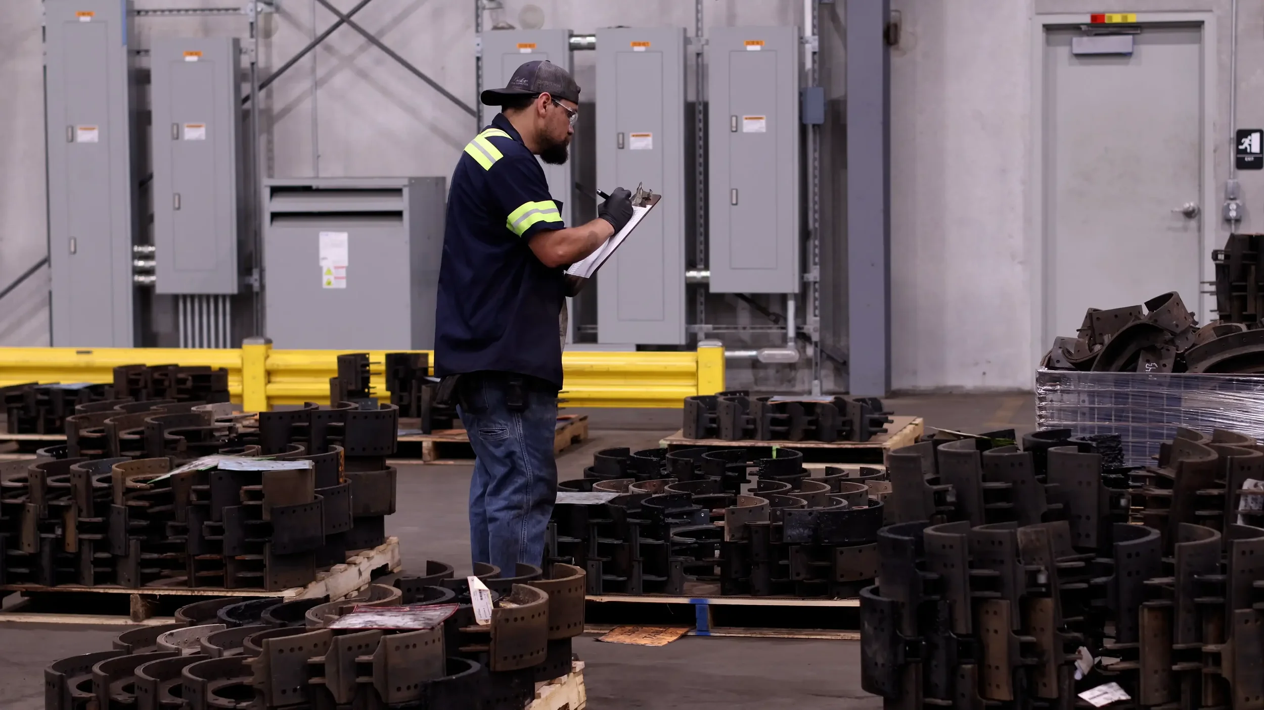 A man in a navy shirt with fluorescent yellow stripes taking notes on a clipboard in an industrial setting with metal parts arranged on pallets.