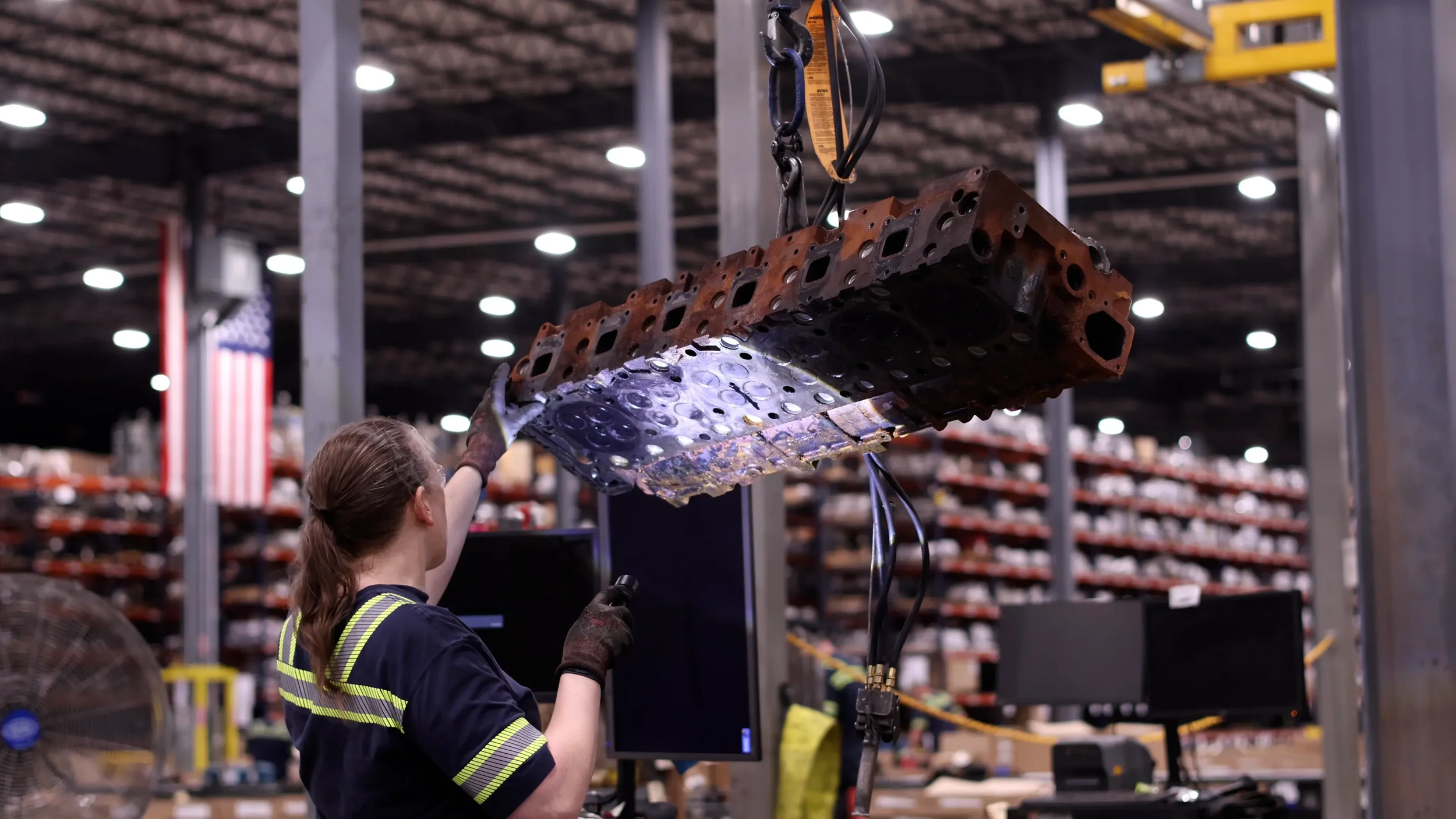 A worker in safety gear handling a large, rusty engine cylinder block with a crane inside an industrial warehouse.