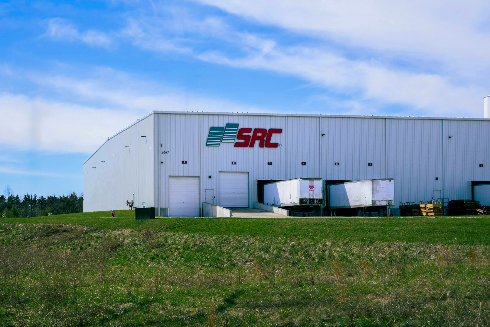 Large white industrial warehouse with the SRC logo on the front, multiple loading docks with trucks, and a grassy field in the foreground under a blue sky with scattered clouds.