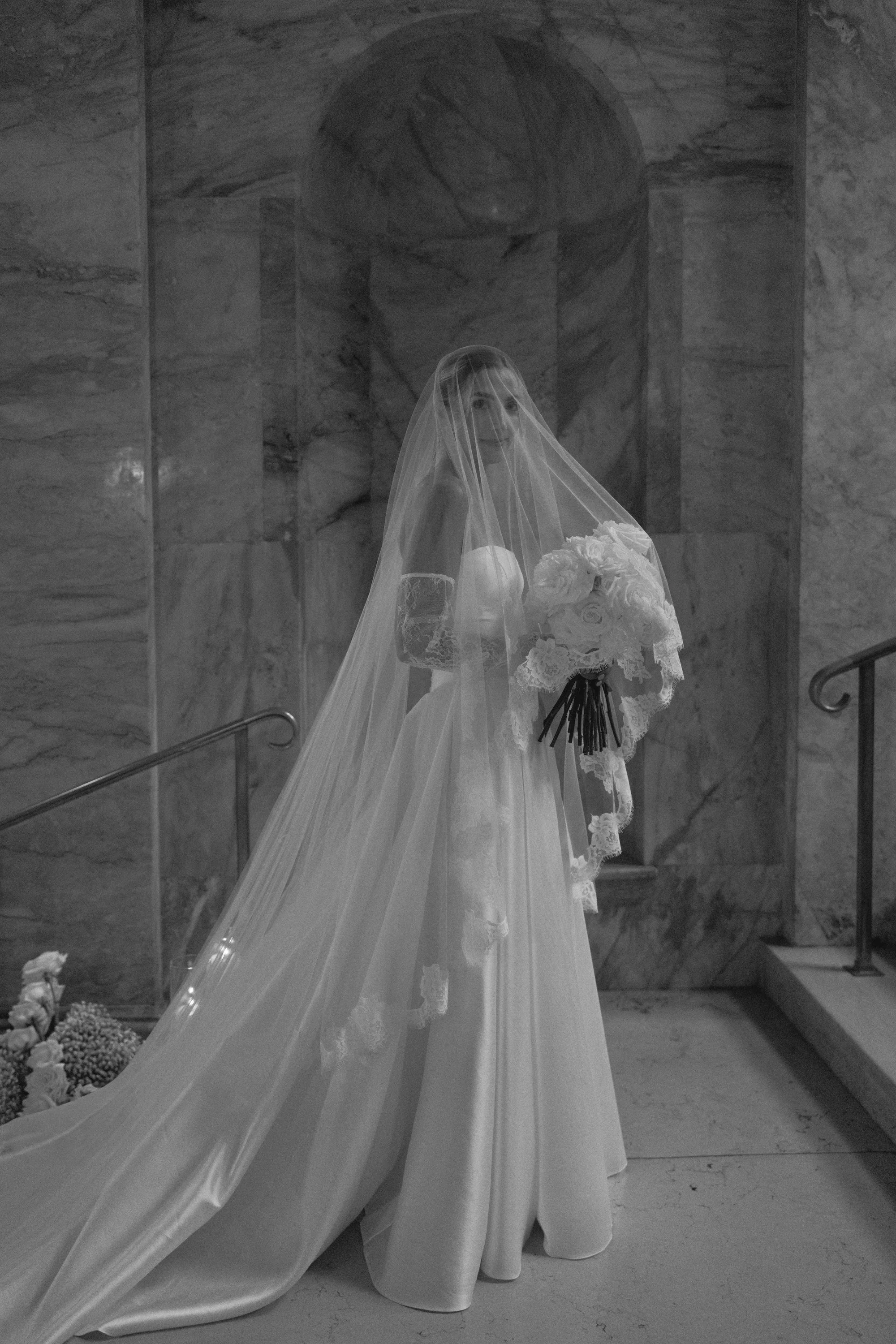 black and white photo of bride looking at camera inside a classy boston downtown venue boston public library with veil over her head holding a bouquet of white flowers