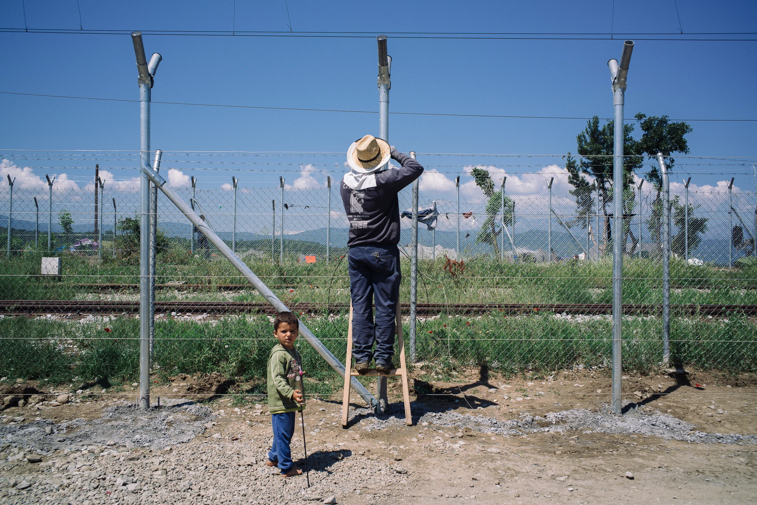 A young Syrian boy innocently watches a man fixing the fence that separates Greece and Macedonia in Idomeni camp.