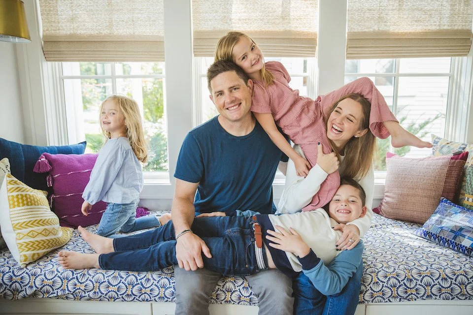 Sophie, her husband, and their three young children sit together with silly, playful smiles. Their son lays across mom and dad's lap, the youngest daughter sits upright on her knees, and the oldest daughter sits across mom and dad's shoulders.
