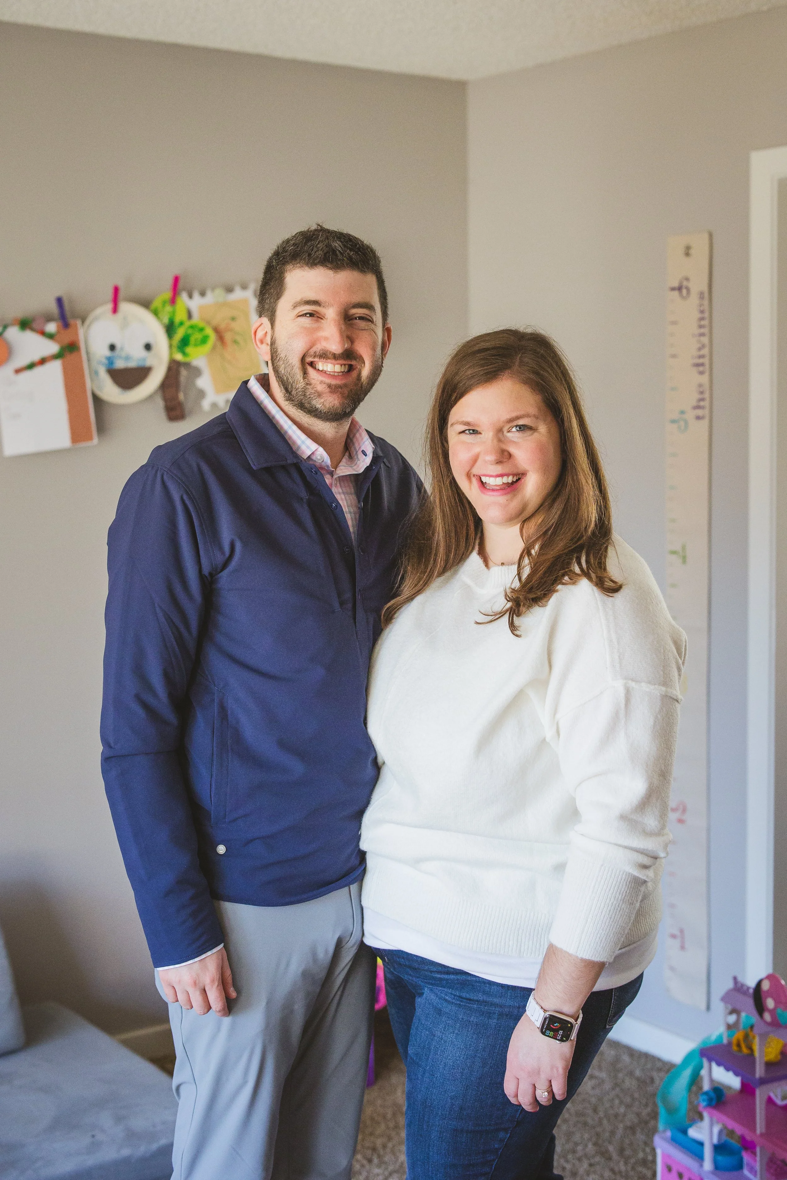 Allie stands with her husband, both smiling warmly in their child's playroom.