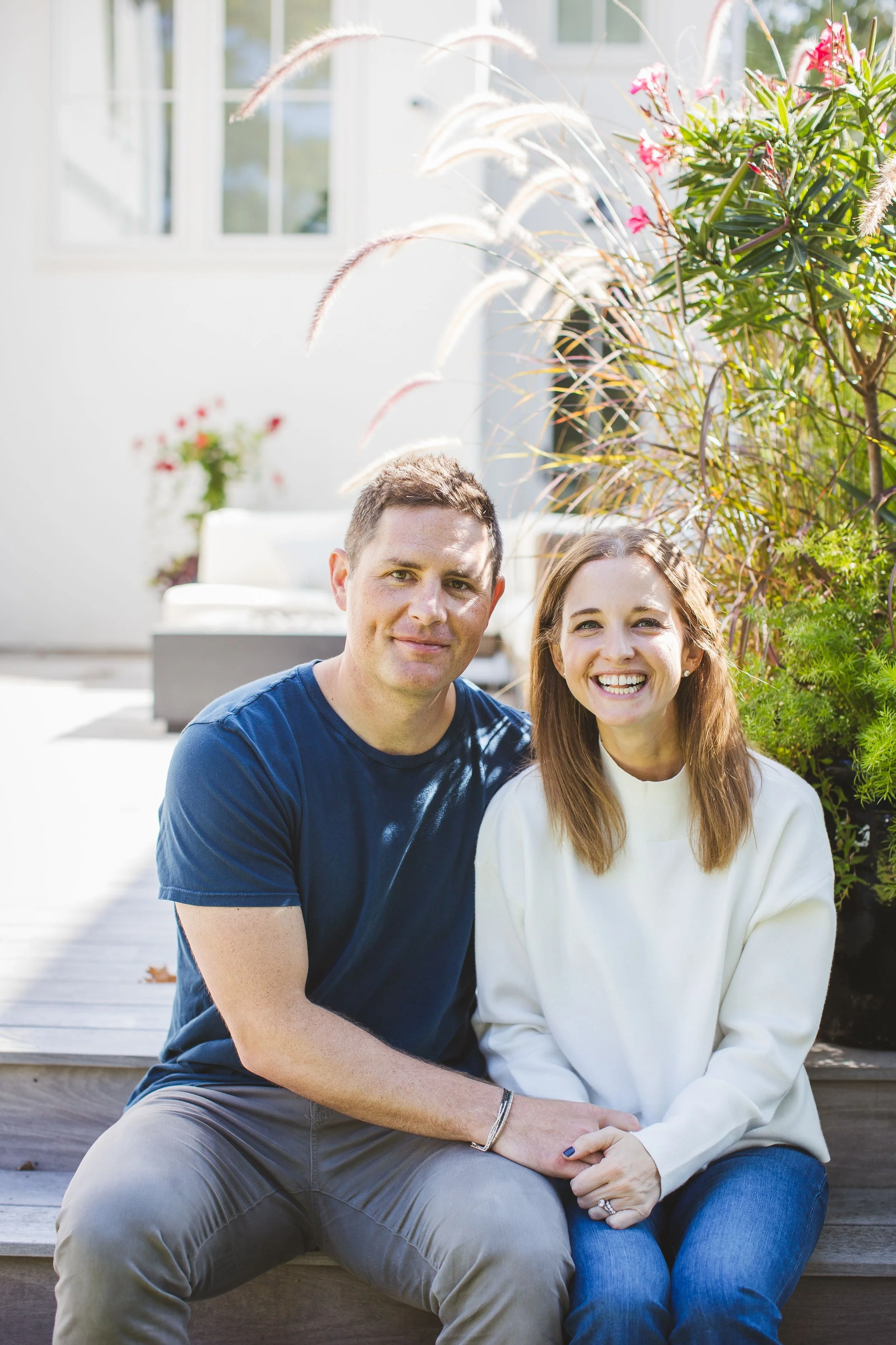 Sophie sits to the right of her husband as they hold hands and smile warmly. They are set against an outdoor backdrop of greenery and pale wood decking.