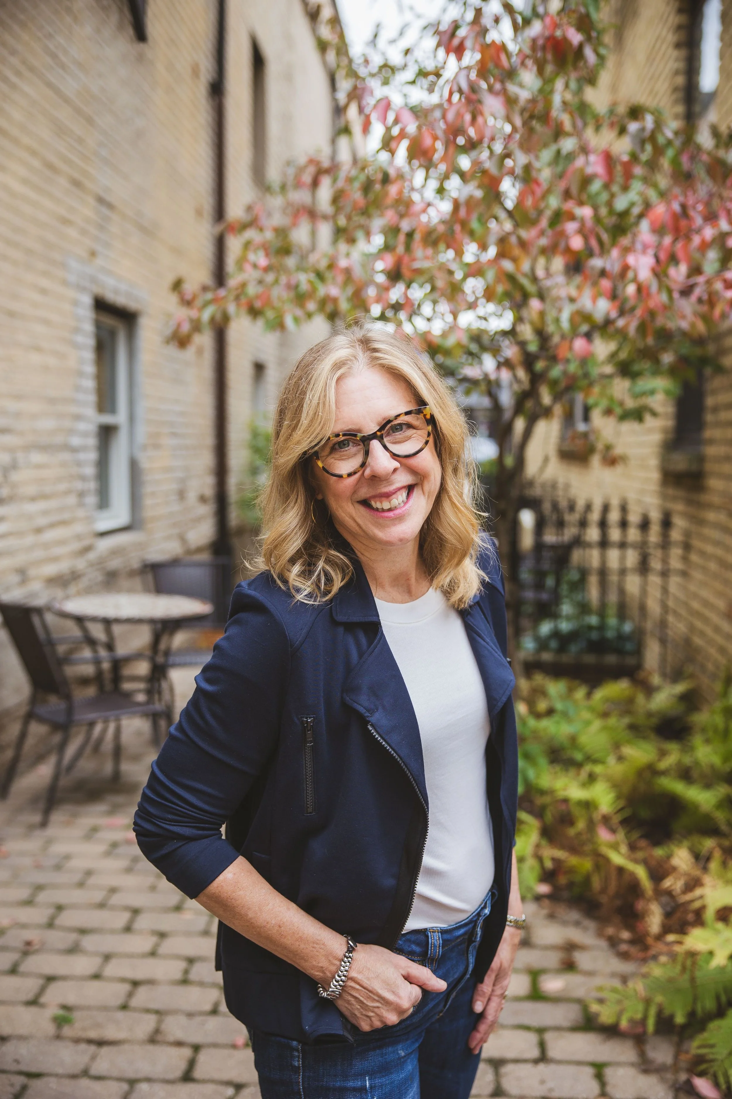 Alison smiles as she poses in a city garden space between two tan brick buildings. She is a middle aged woman wearing glasses and a navy blue jacket.