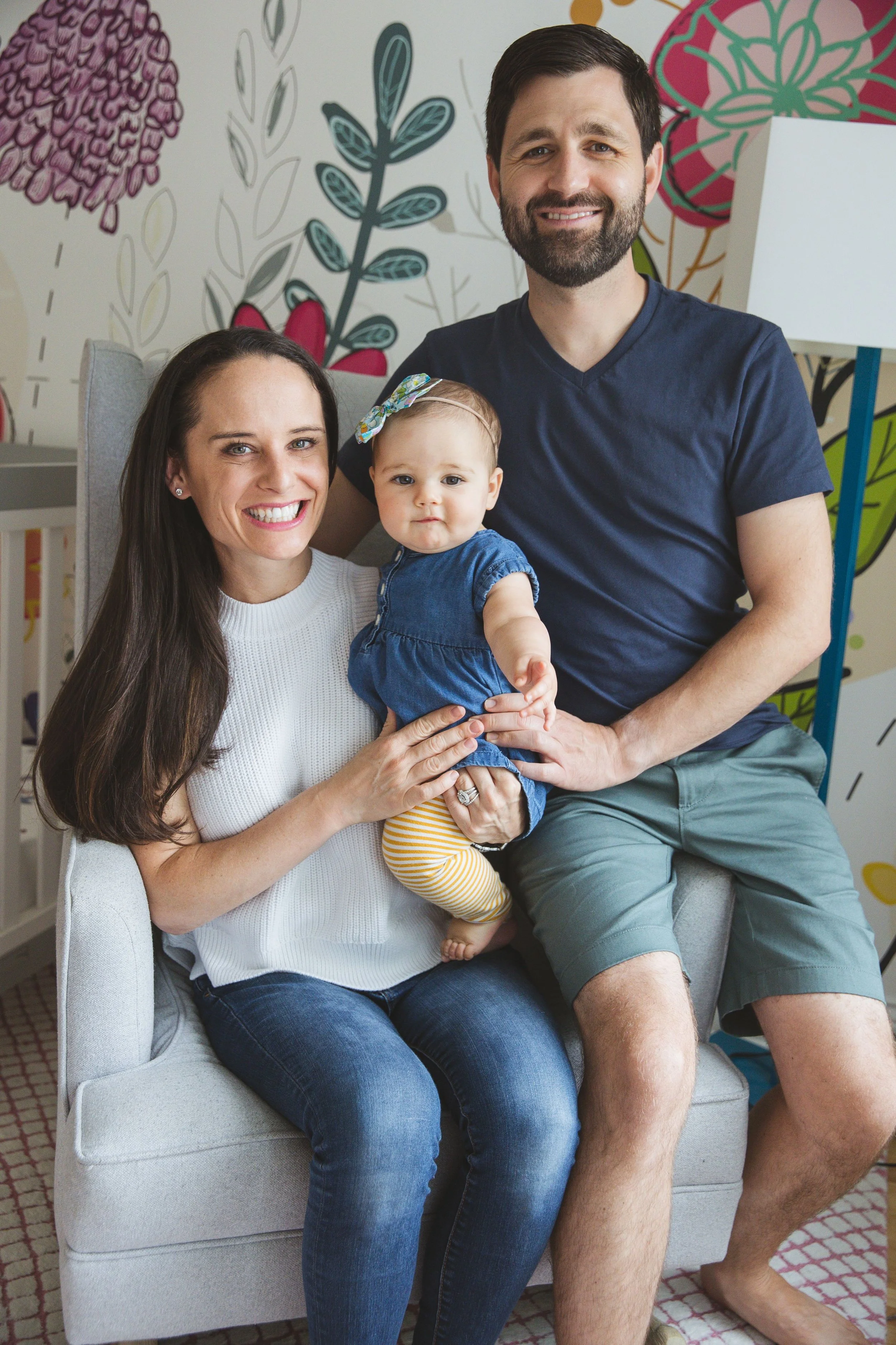Sam holds her baby daughter on her lap while sitting next to her husband in a colorful children's nursery.