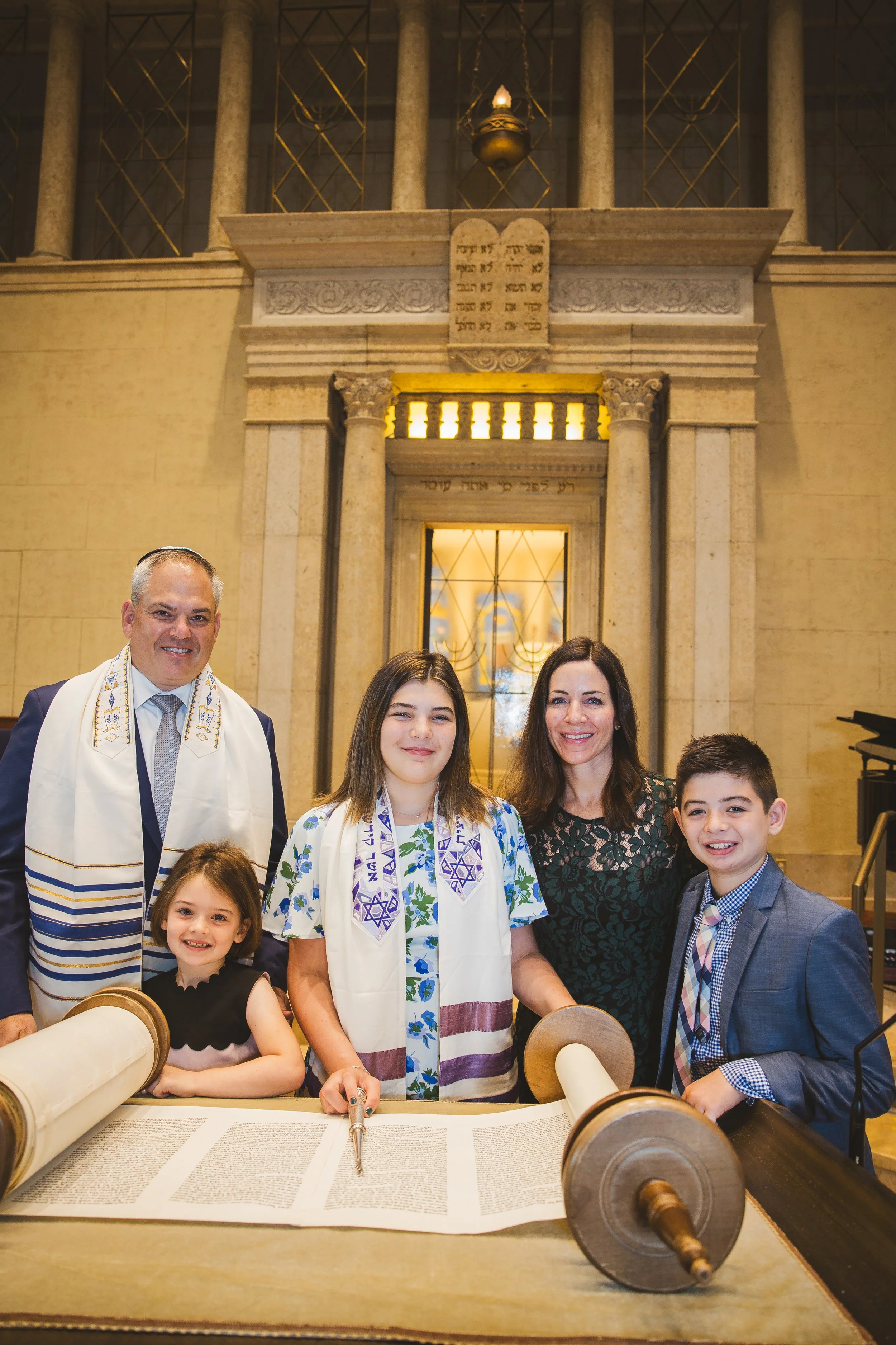 Abby stands with her family as her daughter celebrates her bat mitzvah reads from the Torah. Abby, her husband, and three children stand together smiling in their synagogue in formal suits and dresses.