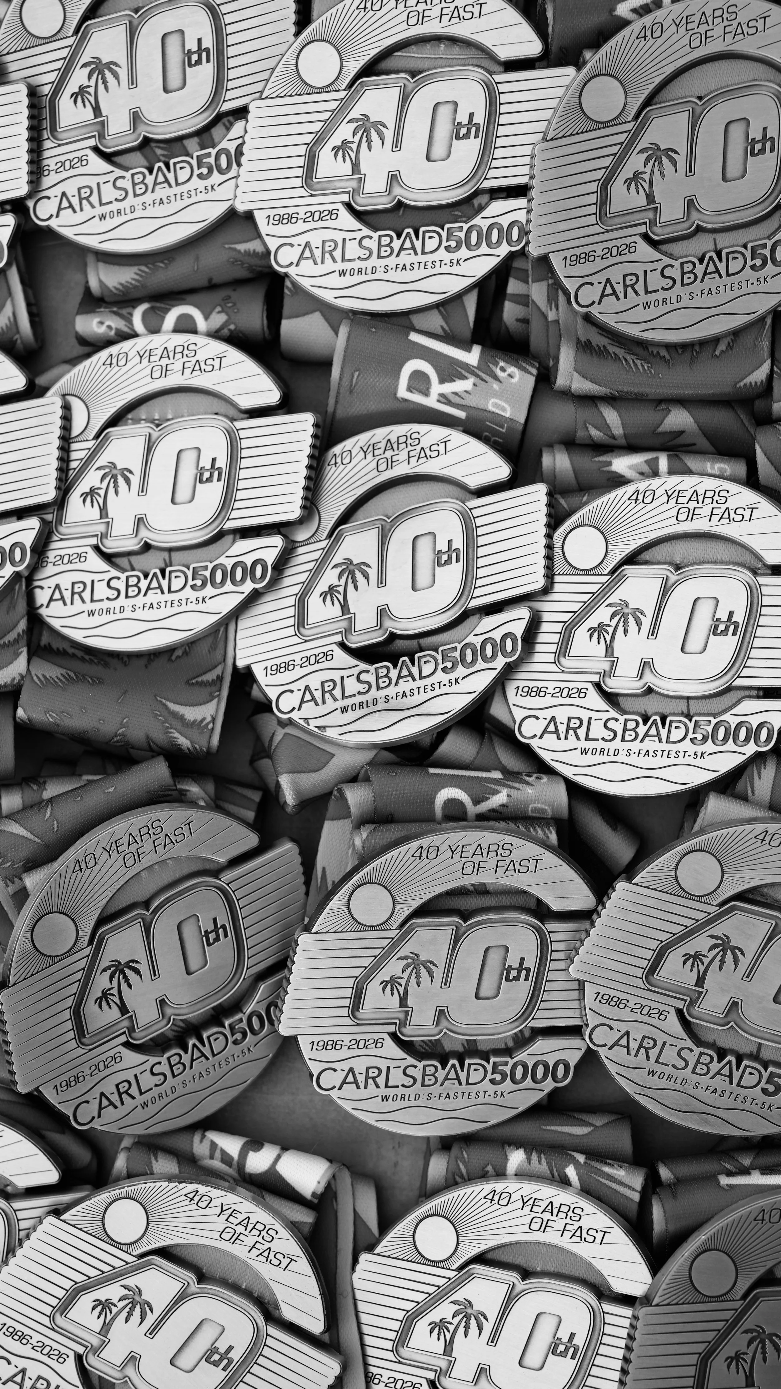 A collection of commemorative medals celebrating 40 years of a marathon race, with the text '40th CARLSBAD 5000, WORLD'S FASTEST 5K,' spanning from 1986 to 2026, featuring palm trees and a sunny design.