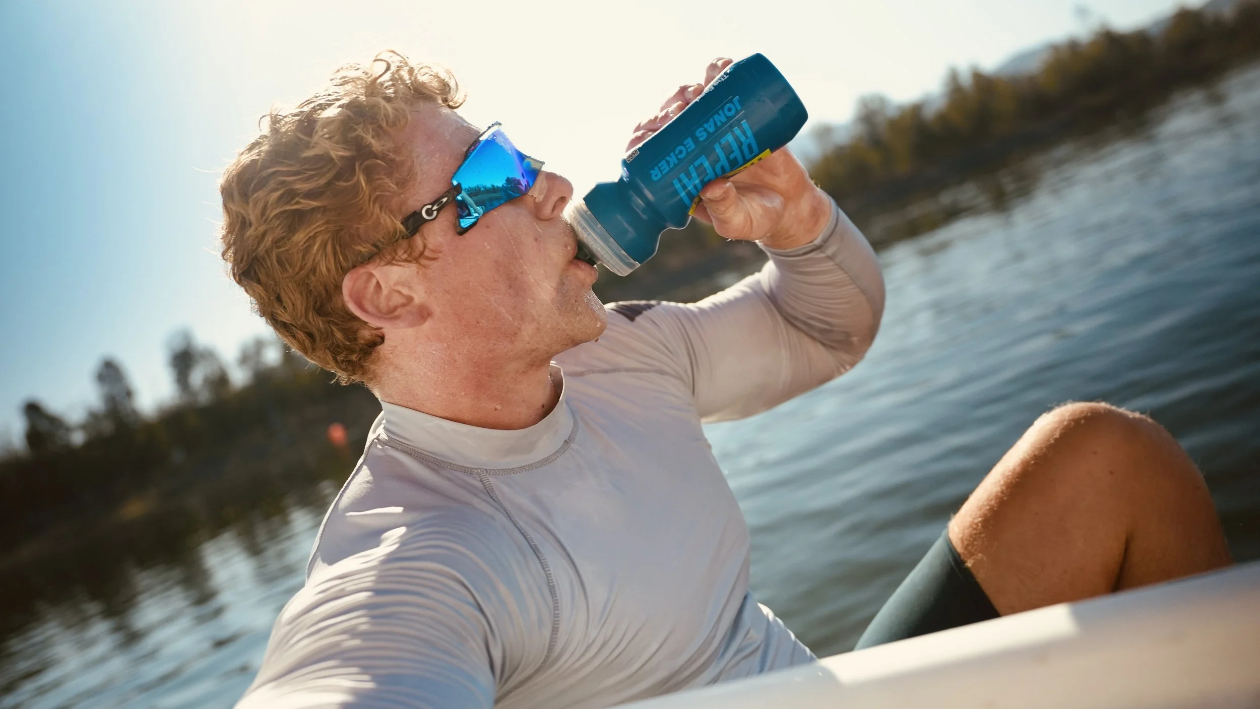 A person with curly curly hair and sunglasses is drinking from a blue sports bottle while sitting by a body of water on a sunny day.