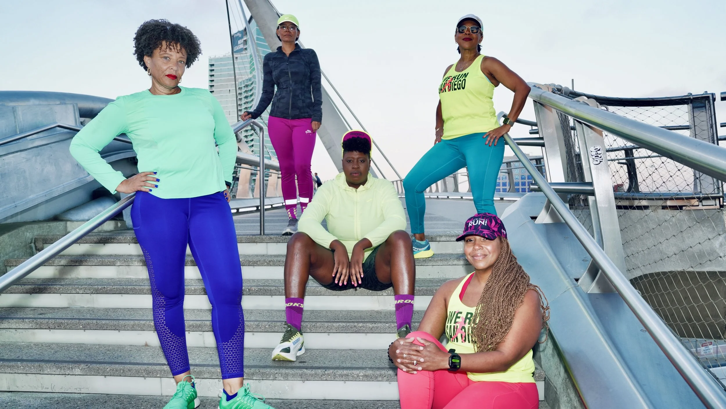 Group of six diverse women dressed in activewear, posing on outdoor stairs with a cityscape and a tall modern building in the background. They appear ready for a workout or run.