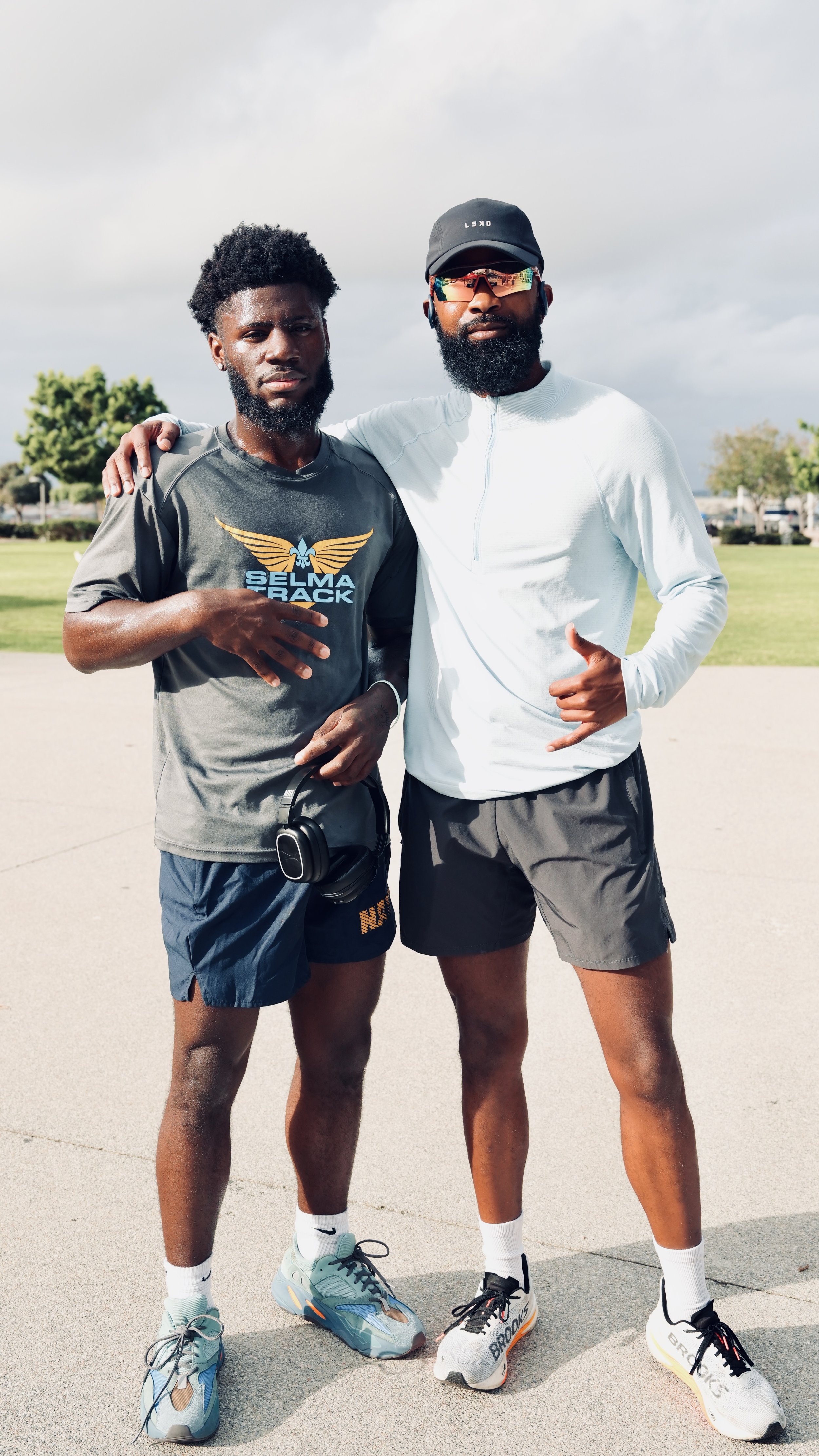 Two men standing outdoors, one with a headphone around his neck, both wearing athletic clothing and sneakers, with their arms around each other's shoulders.