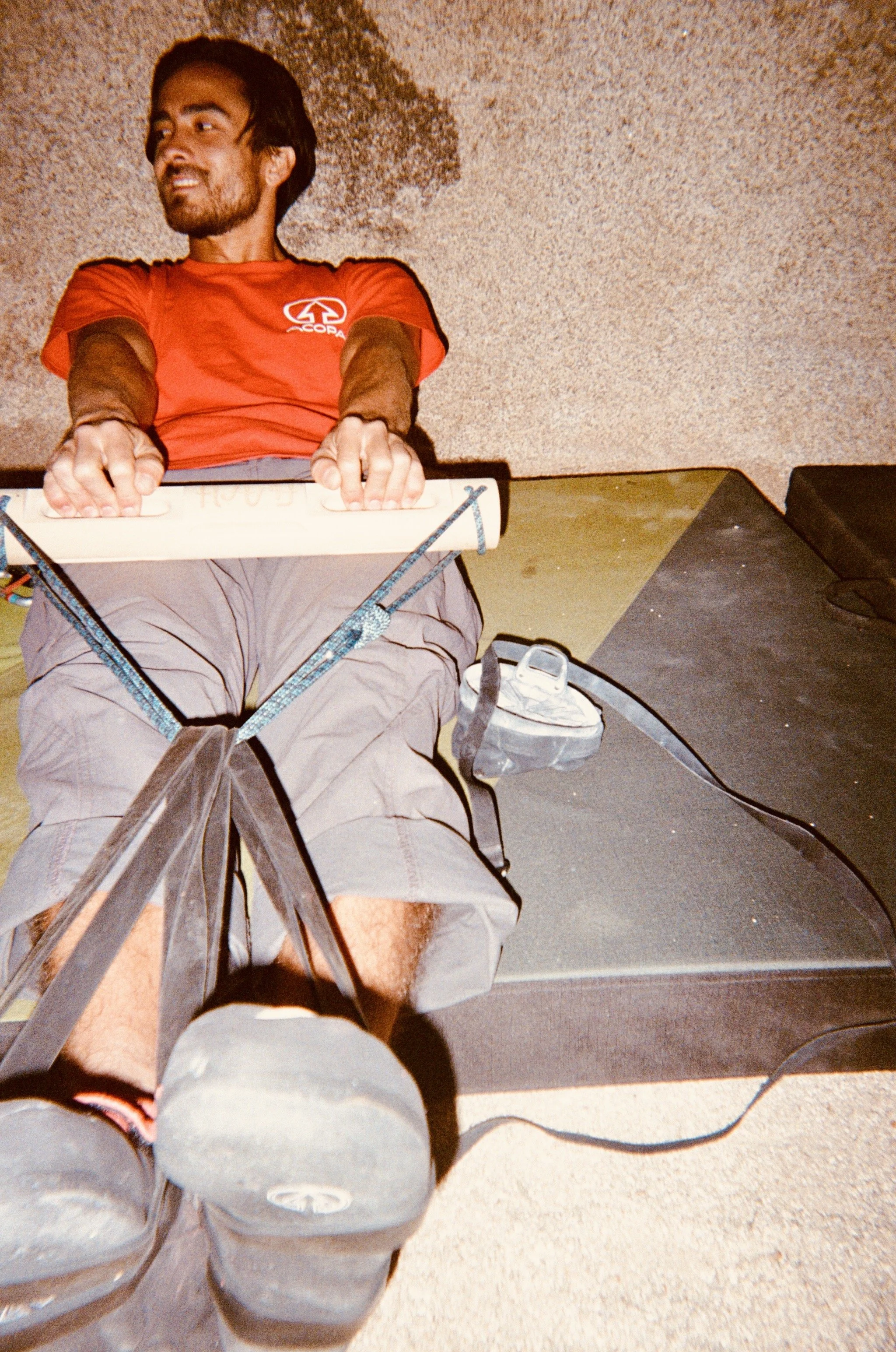A man with a beard and dark hair, wearing a red t-shirt with a white logo, sitting on a chair with his legs extended forward, looking to the side with a smile. The photo appears to be taken from the person's perspective, showing their legs and feet i
