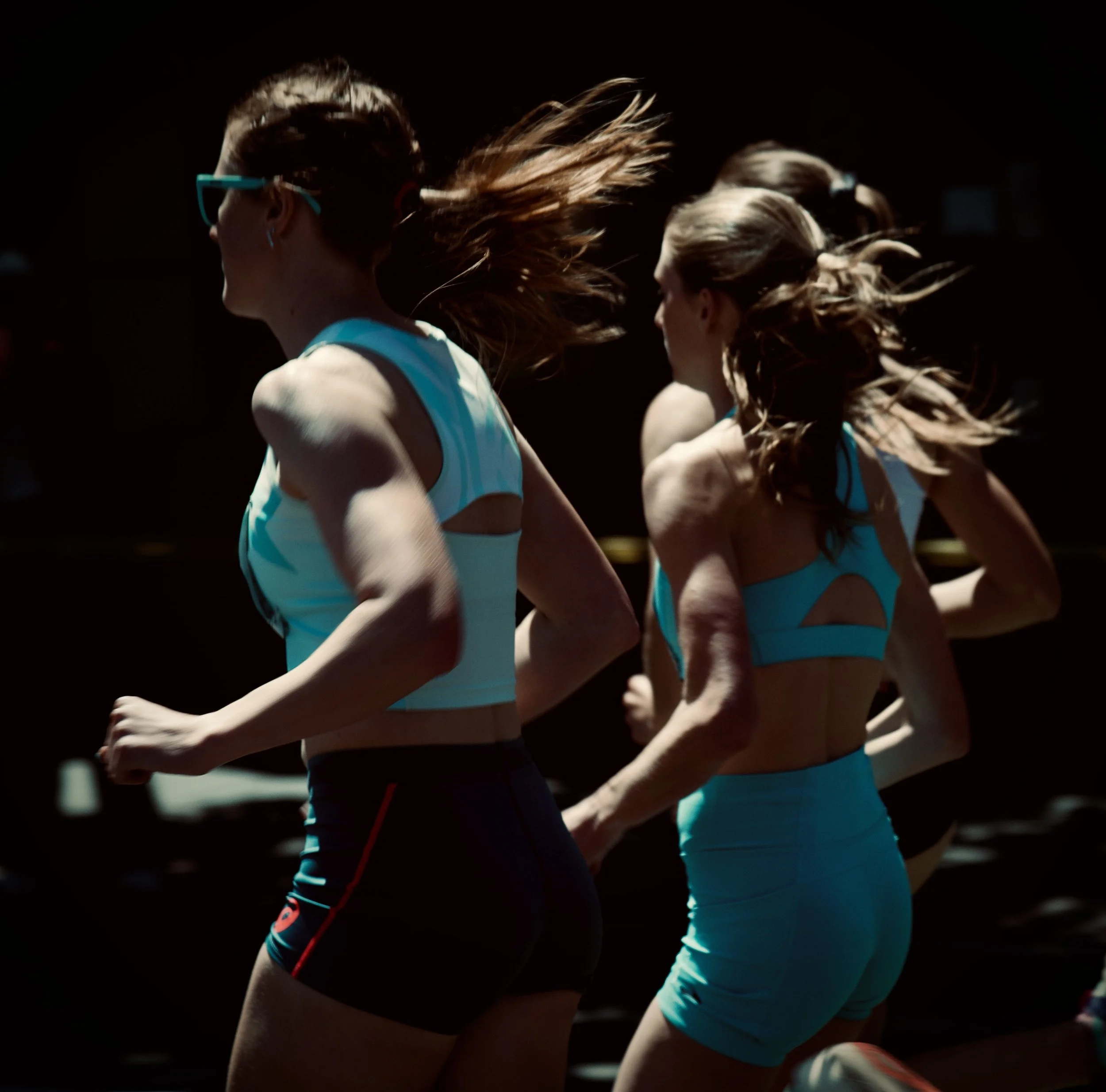 Close-up of female runners in a race, with long hair flowing as they run against a dark background.