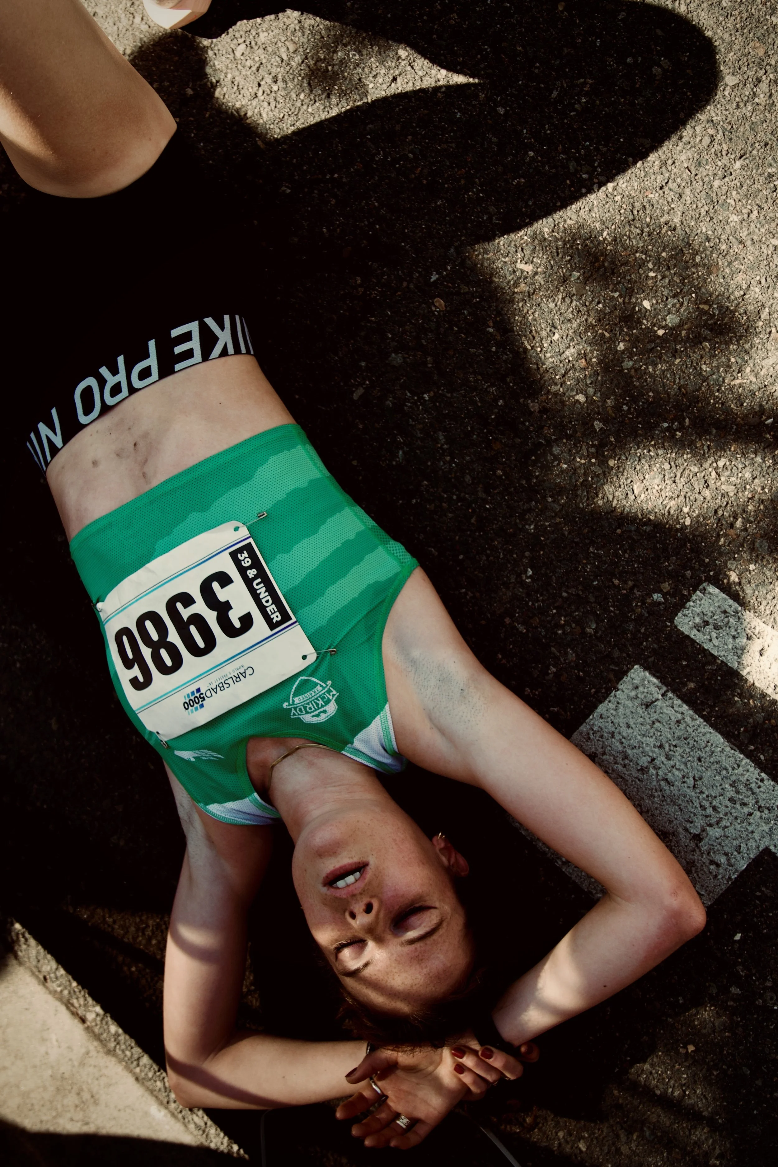 A woman lying on the ground after a race, wearing a race bib with the number 3986, green athletic shorts, and a ring, with her eyes closed and arm resting on her head. The ground is textured and the photo captures her face and upper body.