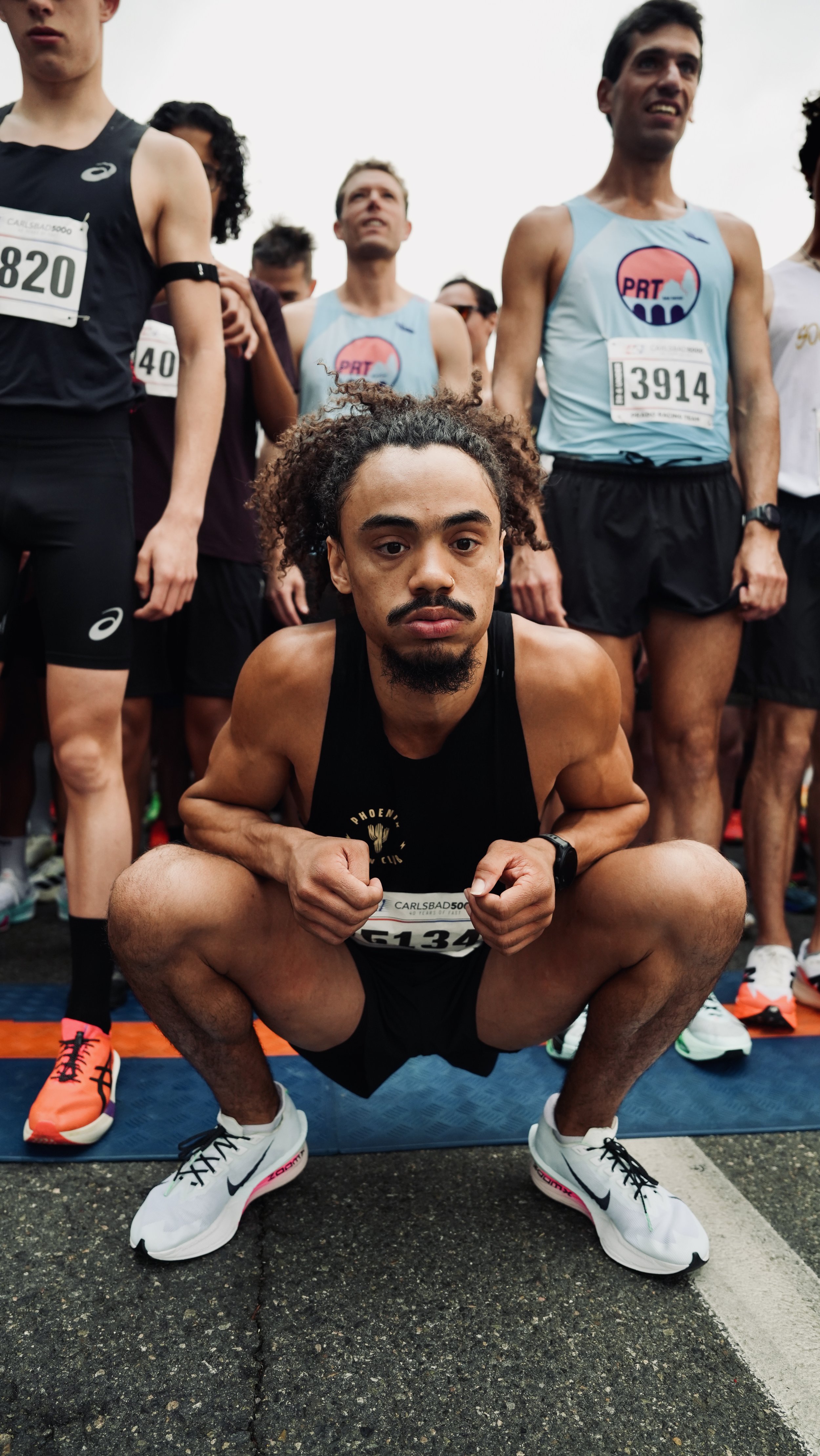A group of marathon runners at the starting line, with one man in the foreground squatting down, wearing white Nike running shoes, a black tank top, and posing with clenched fists. The others are standing behind him, wearing running gear and race bib