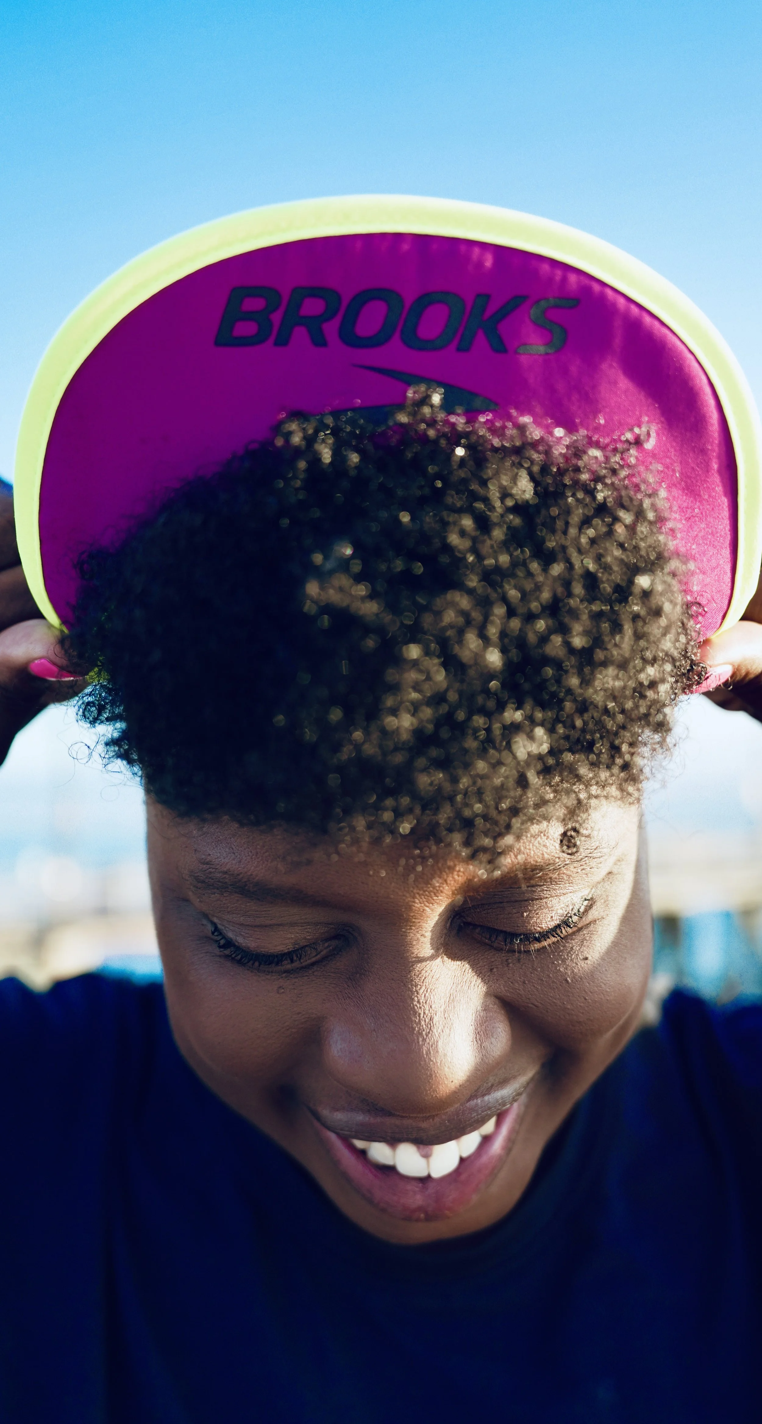 A person with curly hair holding a pink and yellow Brooks-branded frisbee on their head, smiling with eyes closed, outdoors on a sunny day.