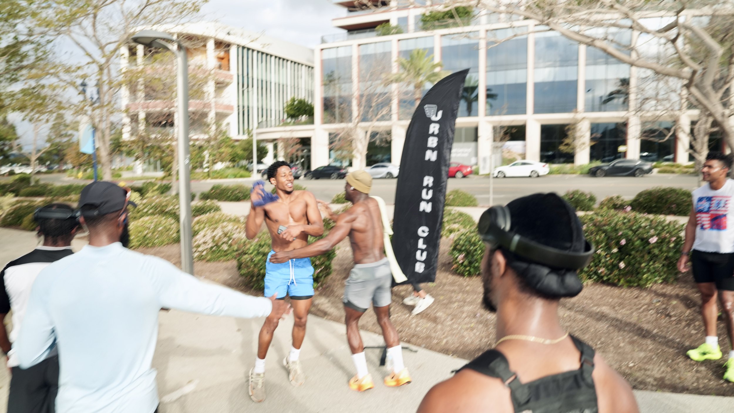 Two shirtless men are standing outside near a landscaped area, hugging and smiling. Several people are observing and taking photos. There is a black flag with white text that reads 'URBAN RUN CLUB,' and modern buildings with glass windows in the back