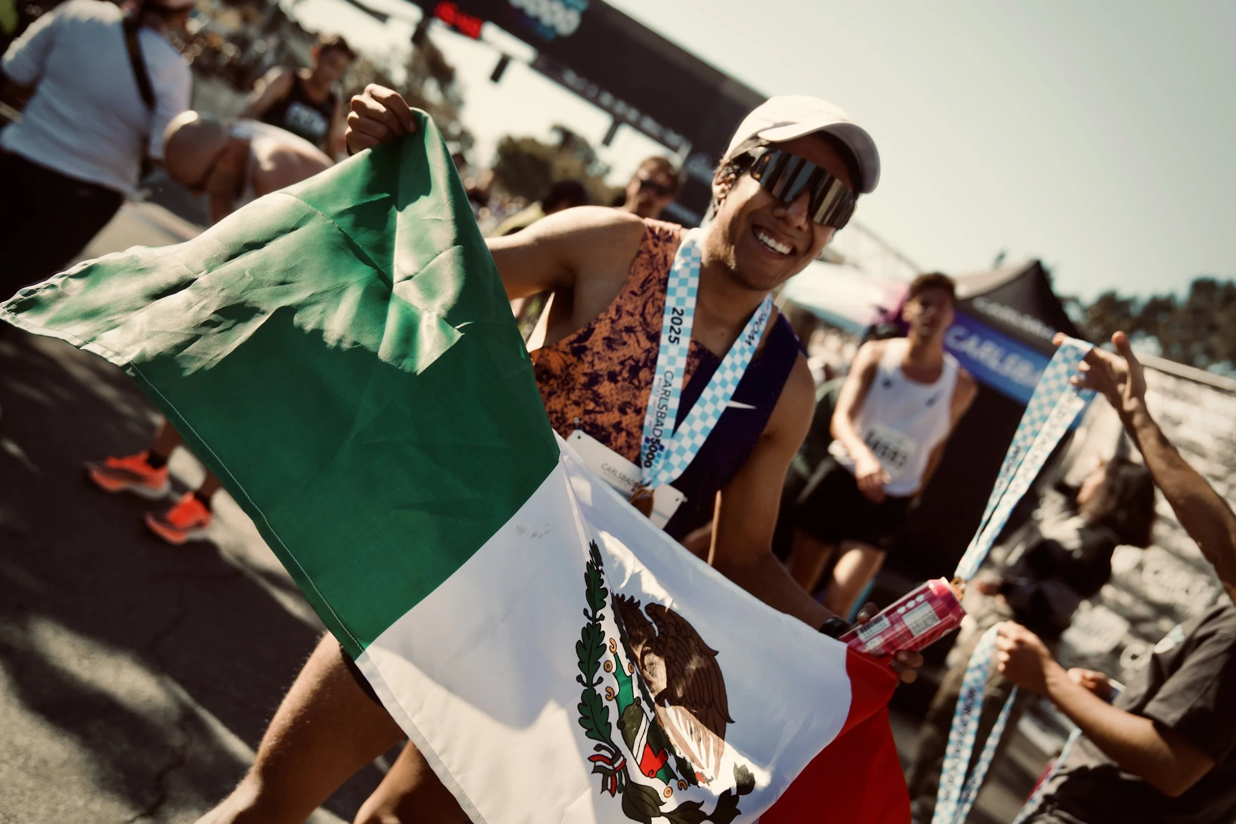 A smiling man wearing sunglasses and a white cap holding a Mexican flag at an outdoor event, with other people and event banners visible in the background.