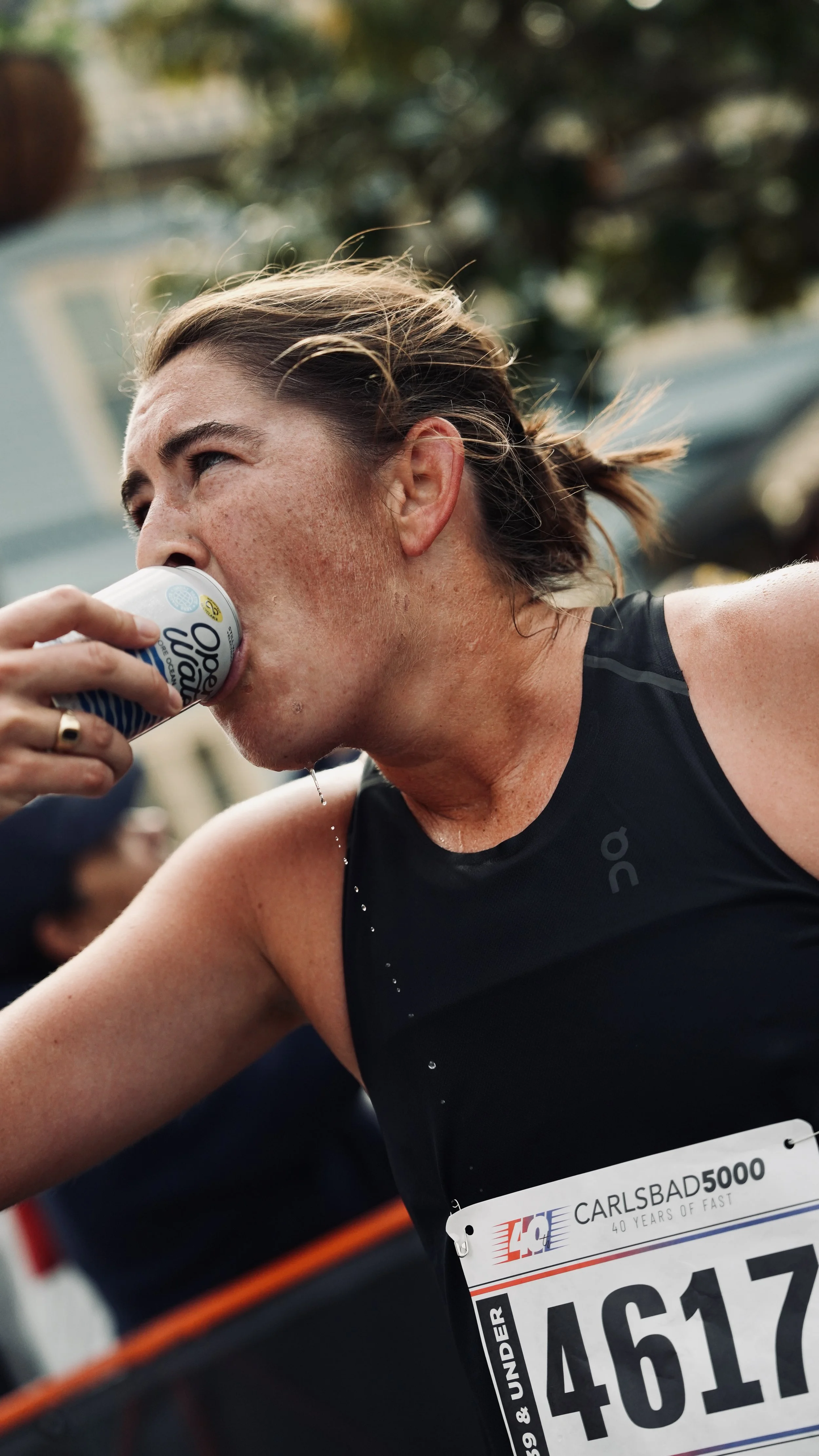 A female marathon runner sipping water during a race, wearing a black athletic top with a race bib number 4617 from the Carlsbad 5000 race, with trees and race barriers in the background.