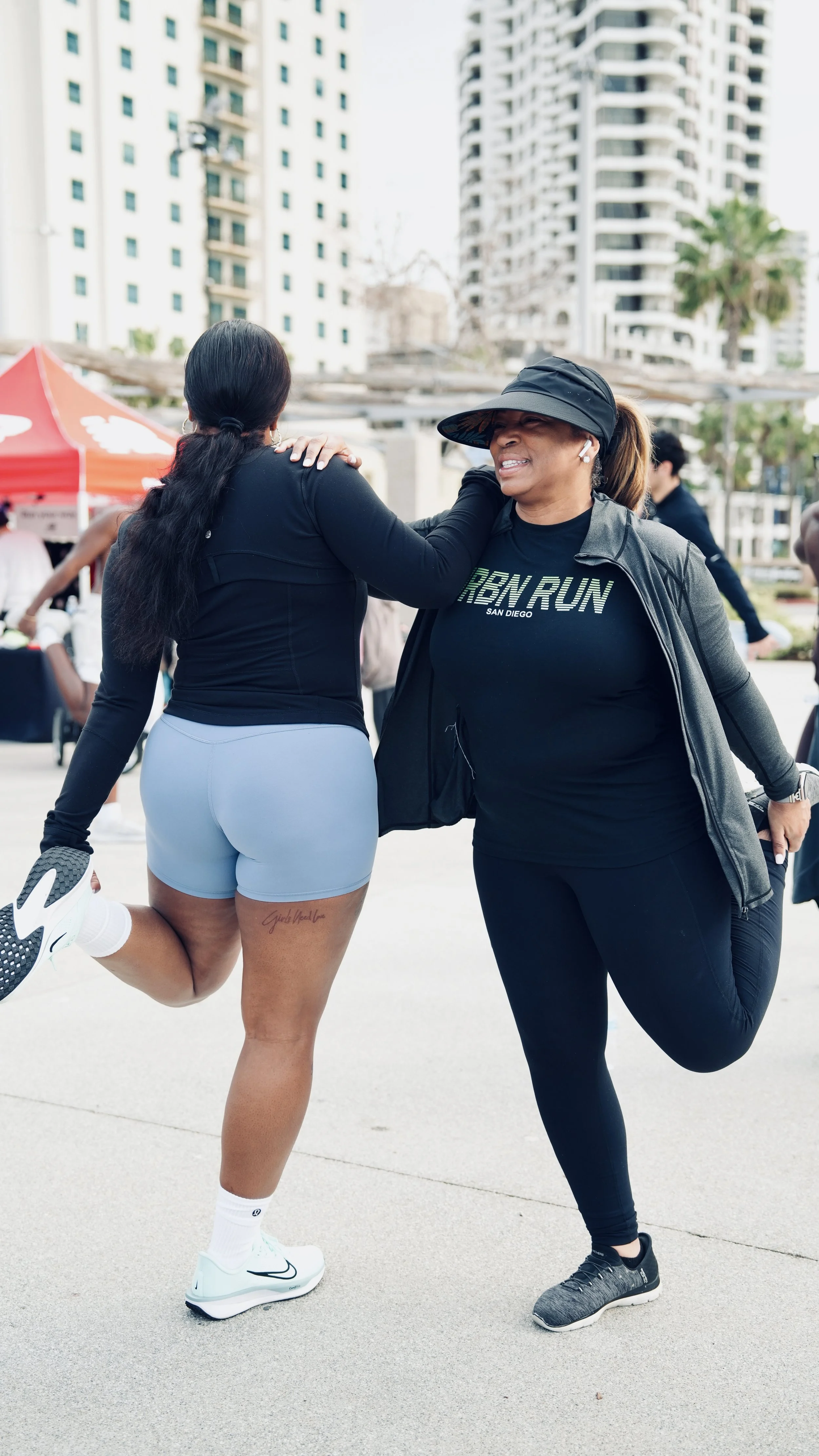 Two women smiling and embracing during an outdoor event with high-rise buildings in the background. One woman is in workout clothes, holding a helmet, and the other woman is in black athletic wear with a visor, both with one leg raised behind them.