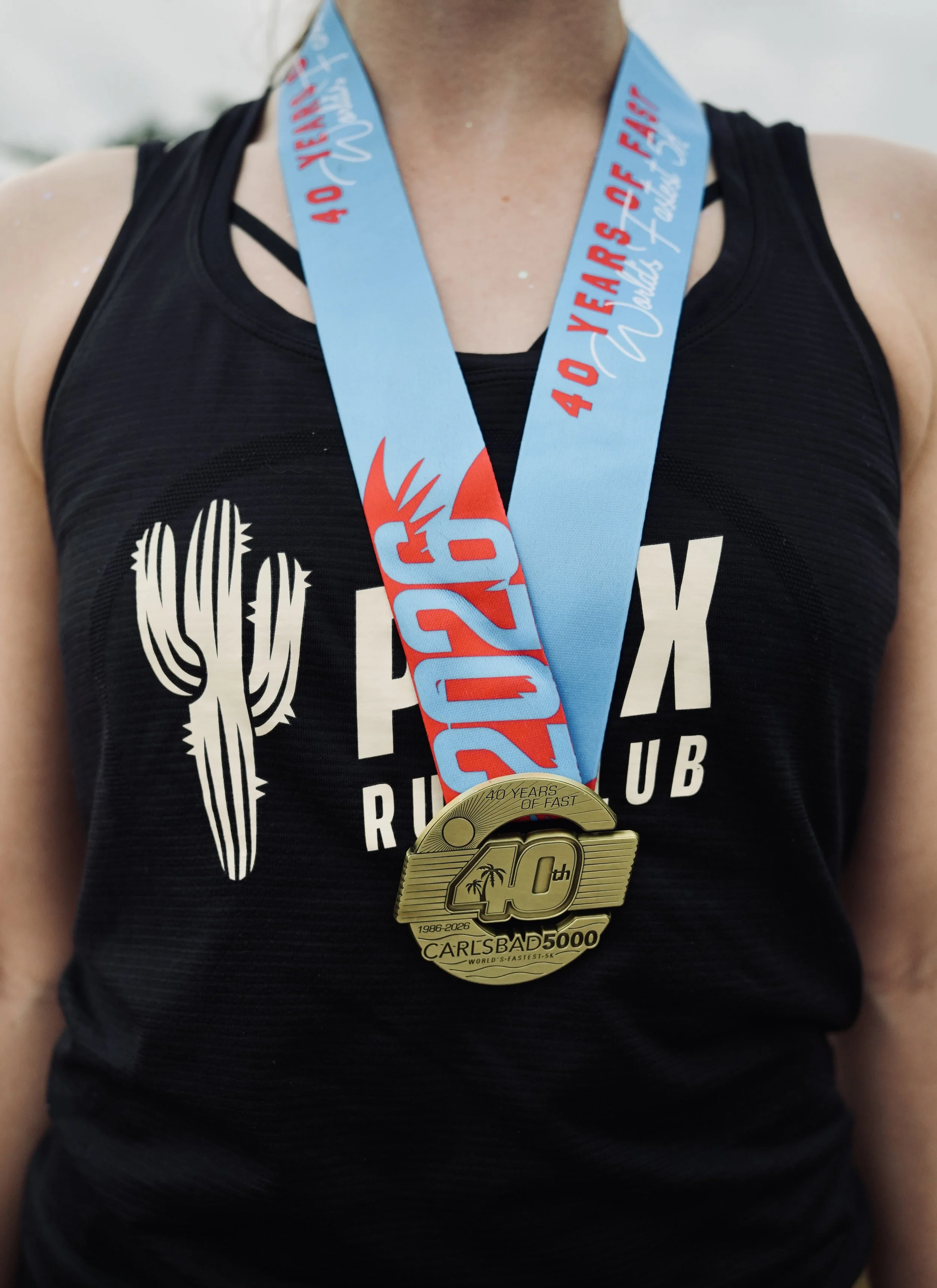 Close-up of a person wearing a black tank top with a graphic design, and a gold medal with a ribbon around their neck, celebrating a running race achievement.