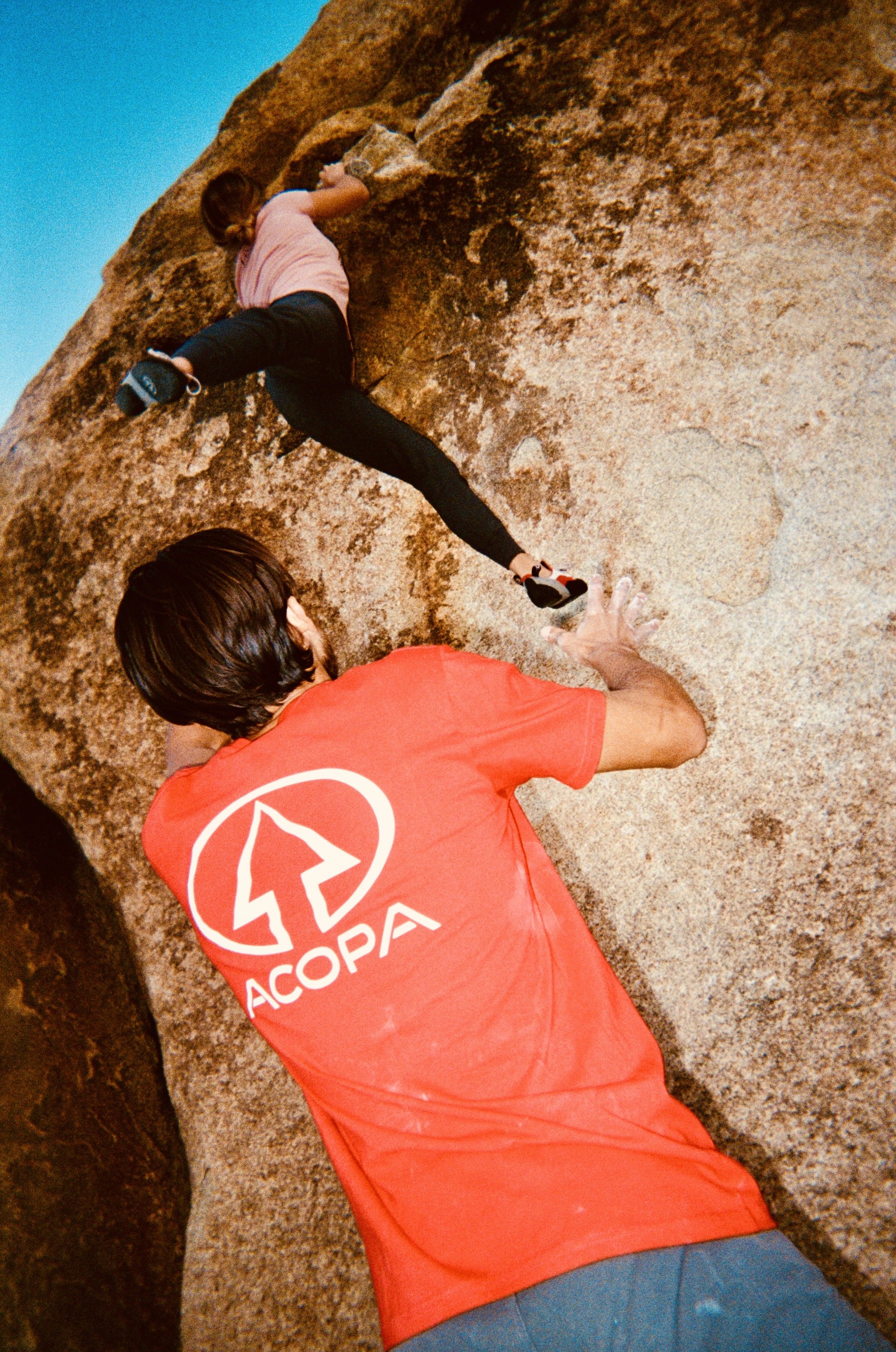 A person wearing a red shirt with a white logo and the word 'ACOPA' on the back is bouldering on a large rock formation. They are reaching with their left hand for a hold, while their right hand and feet are on the rock.