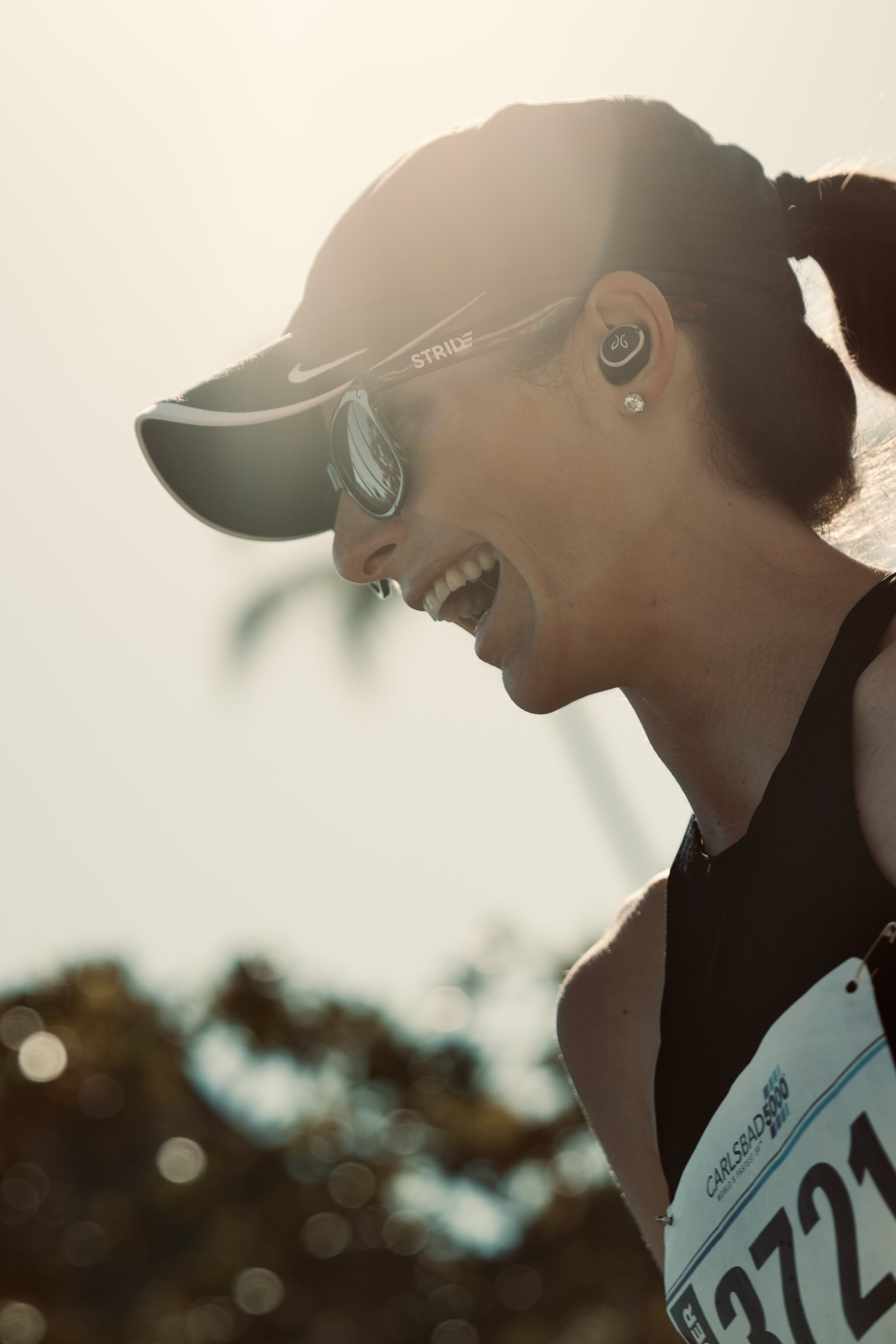 A woman smiling, wearing a black sports hat, sports glasses, wireless earbuds, and a race bib, outdoors with blurred trees and bright sunlight.