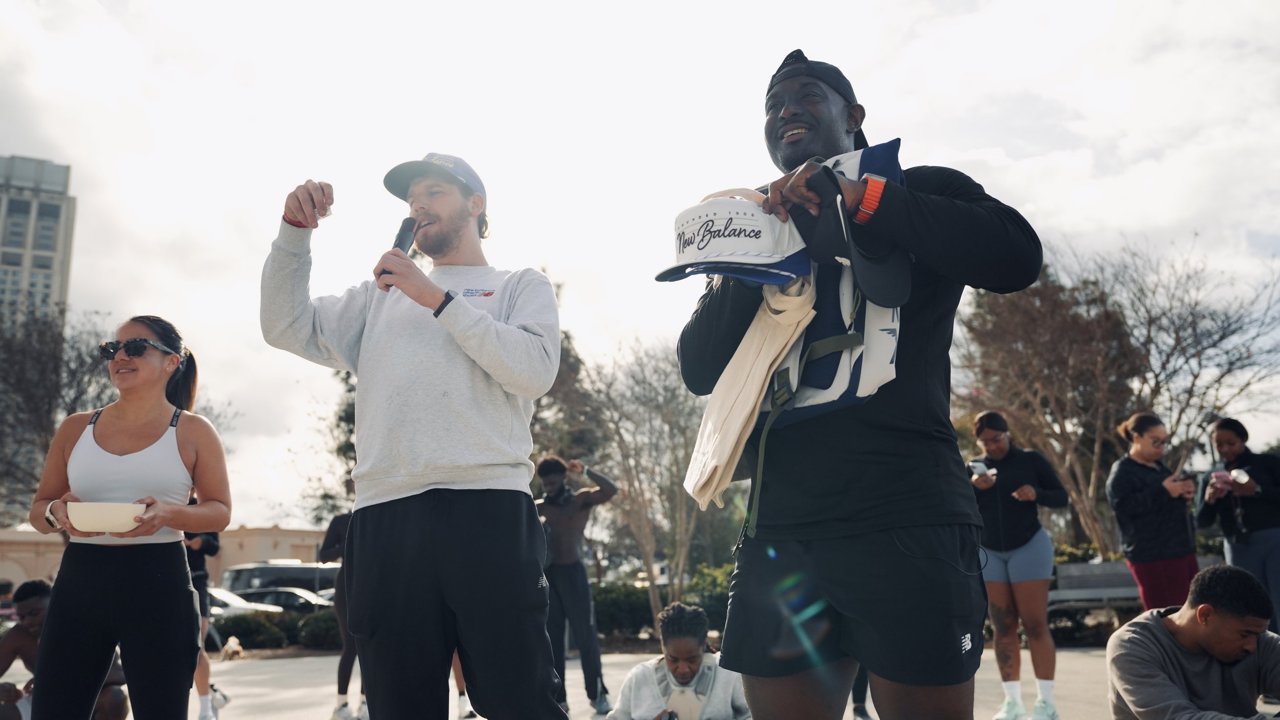 A group of people gathered outdoors, some standing and some sitting, with two men in the foreground, one speaking into a microphone and holding a helmet, and a woman wearing sunglasses holding a bowl, with others nearby using their phones.