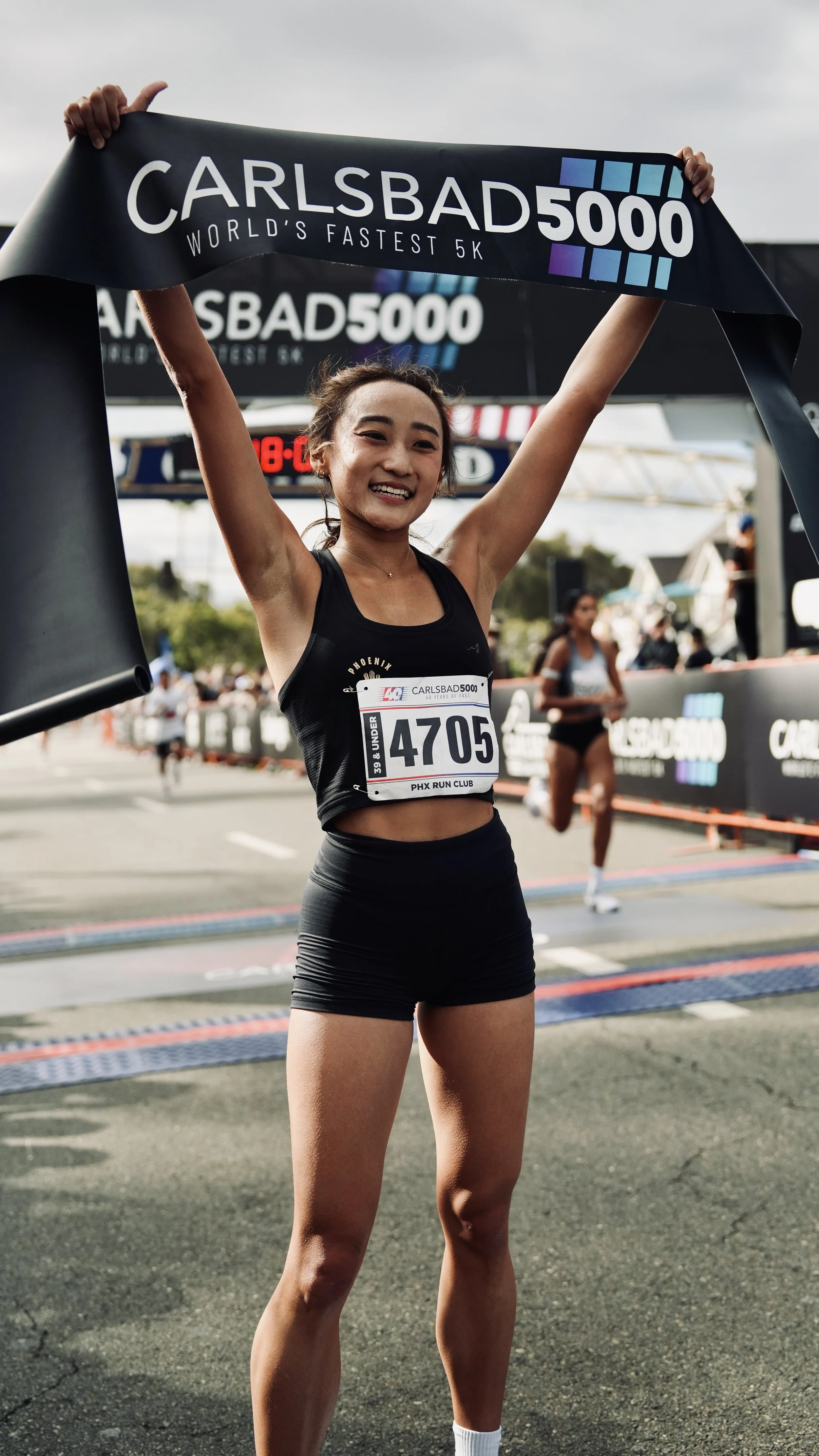 A young female runner crossing the finish line at the Carlsbad 5000 race, smiling and holding up the event banner, with other runners and race signage in the background.