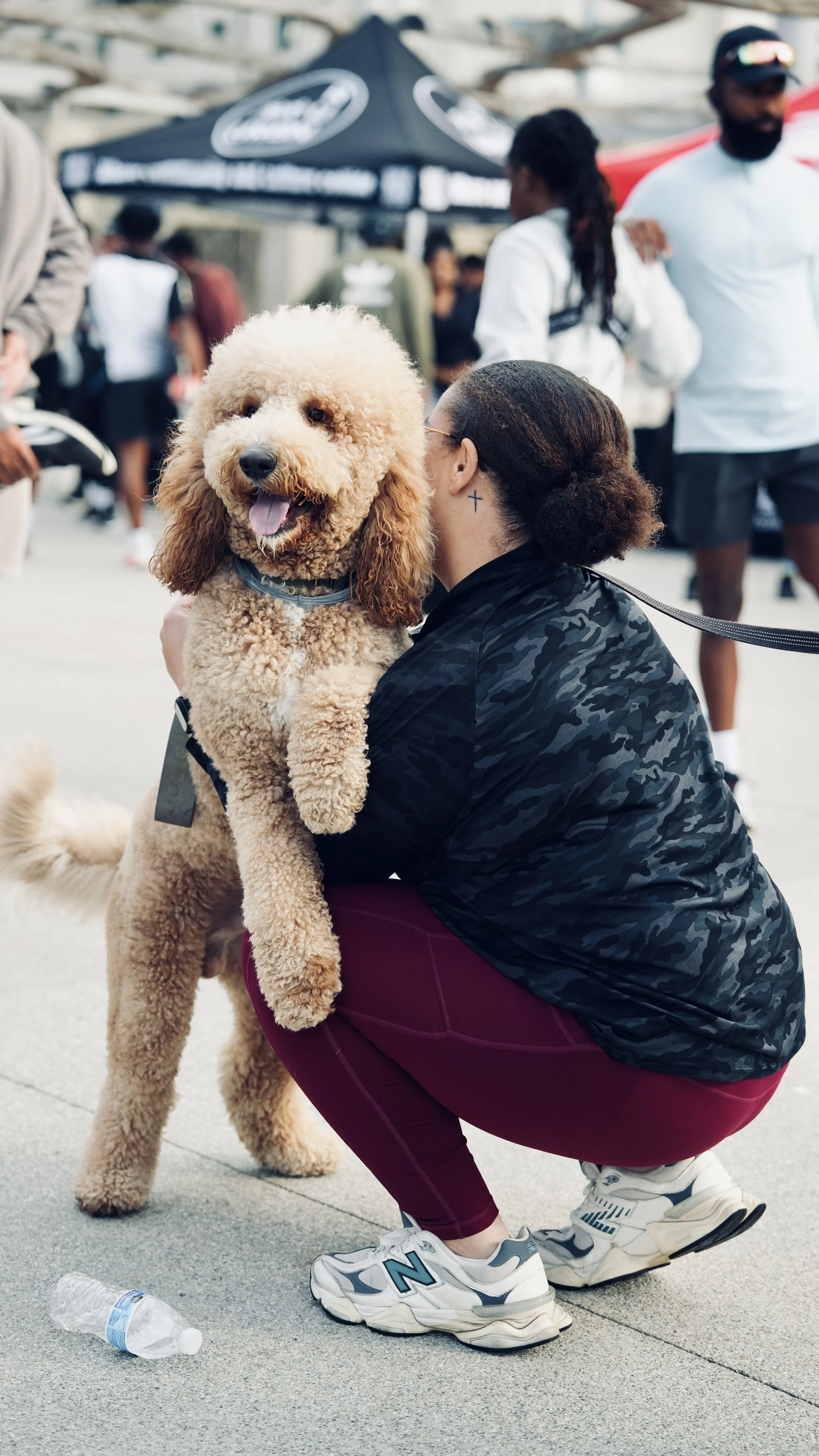 A woman with dark hair tied in a bun and a tattoo behind her ear, wearing a black camo jacket and maroon pants, crouched down holding a happy, curly-haired goldendoodle dog with a blue collar, in an outdoor event with blurred people in the background