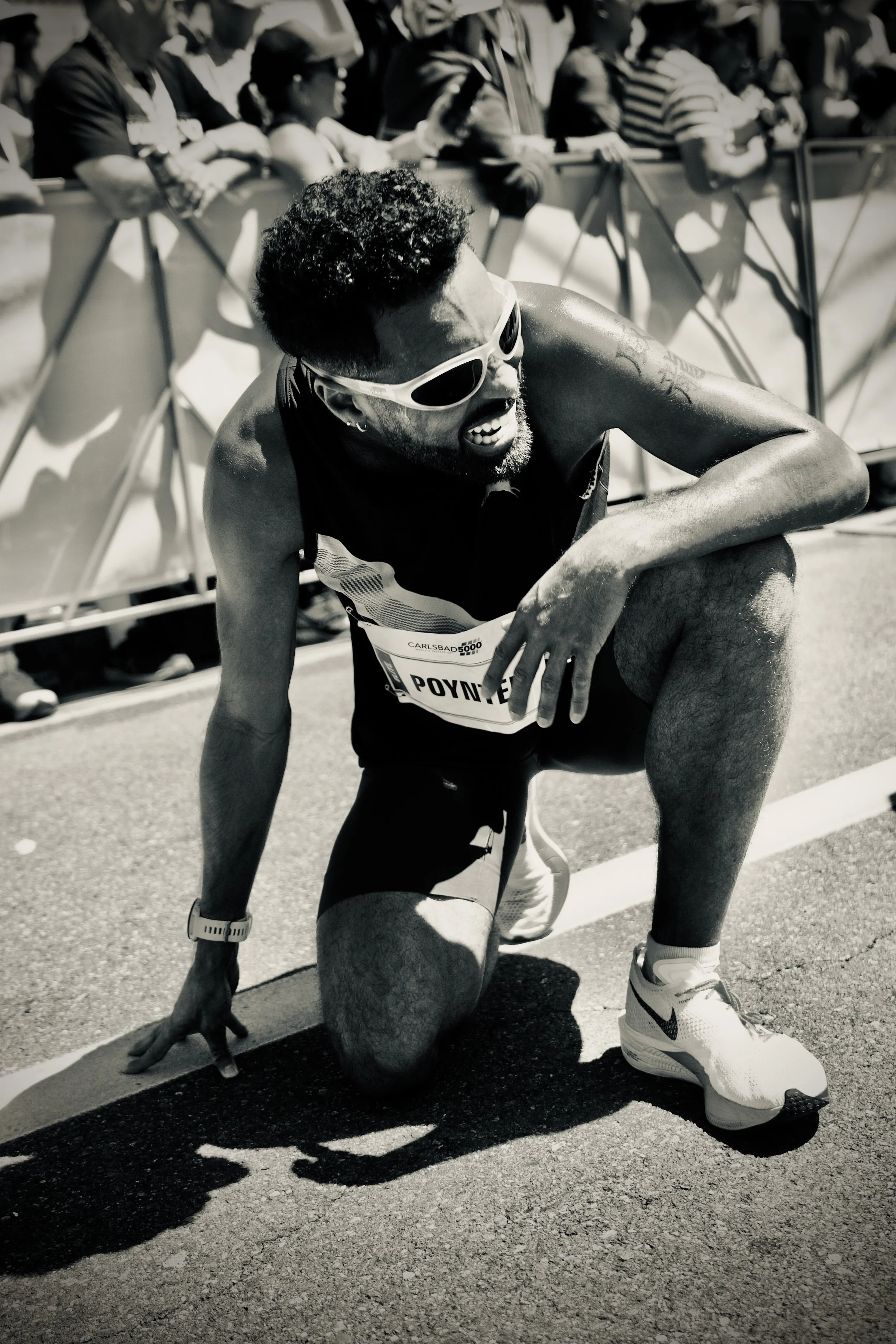 A male runner sitting on the ground, holding his knee, wearing sunglasses, shorts, and athletic shoes, with a race bib labeled 'POYNTER'. He appears to be in pain after a race, with spectators behind a barrier watching.