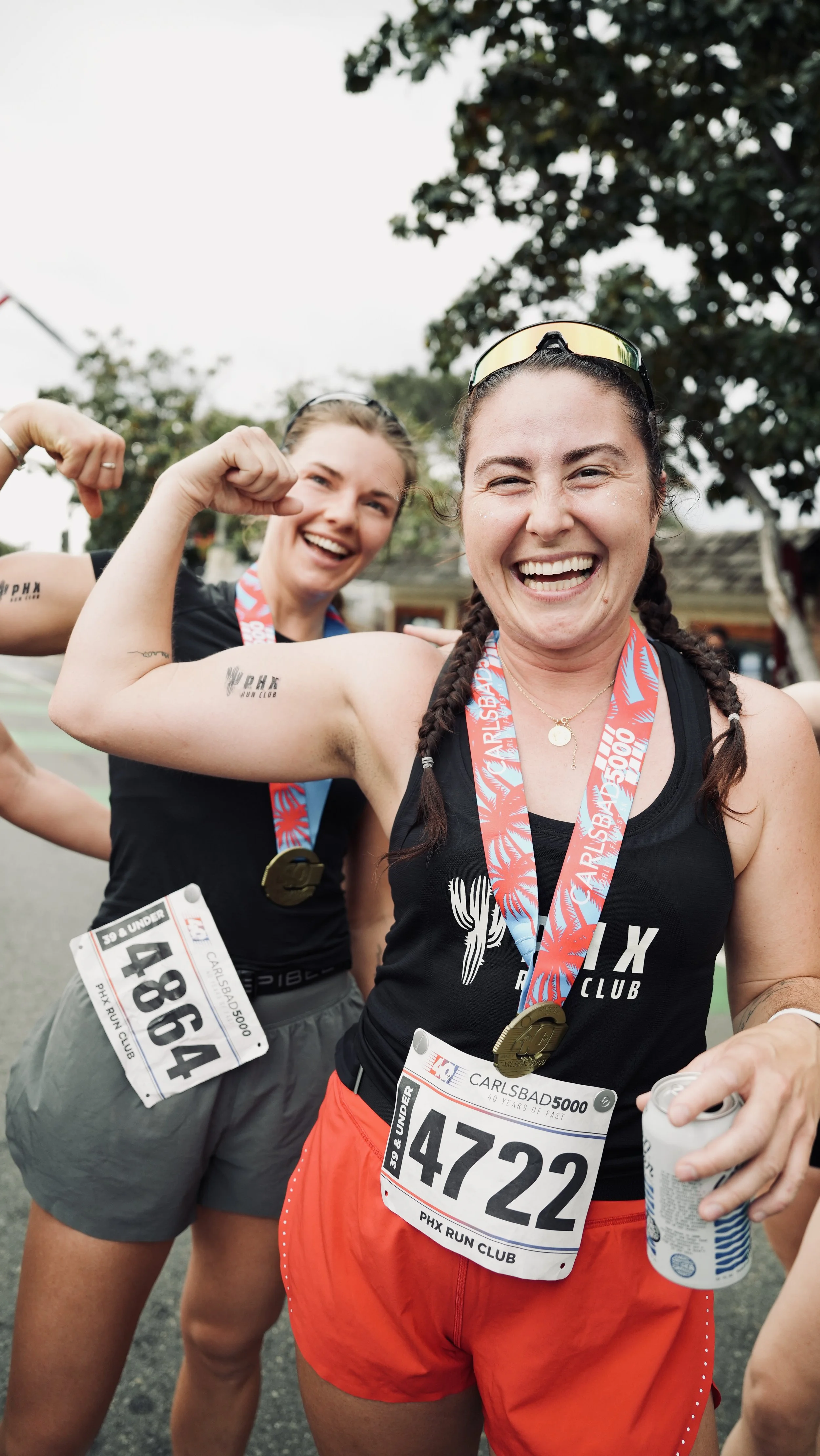 Two women smiling, flexing their biceps, wearing race bibs and medals after finishing a race outdoors.