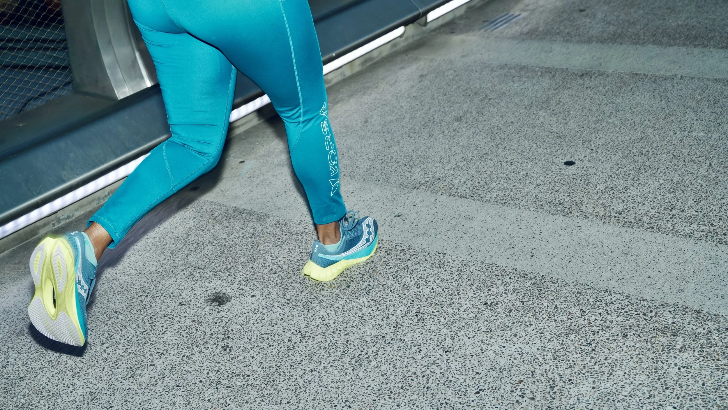 A person wearing blue athletic leggings and running shoes kneeling on a gray textured sidewalk near a metal bench with a greenish electrical box in the background.