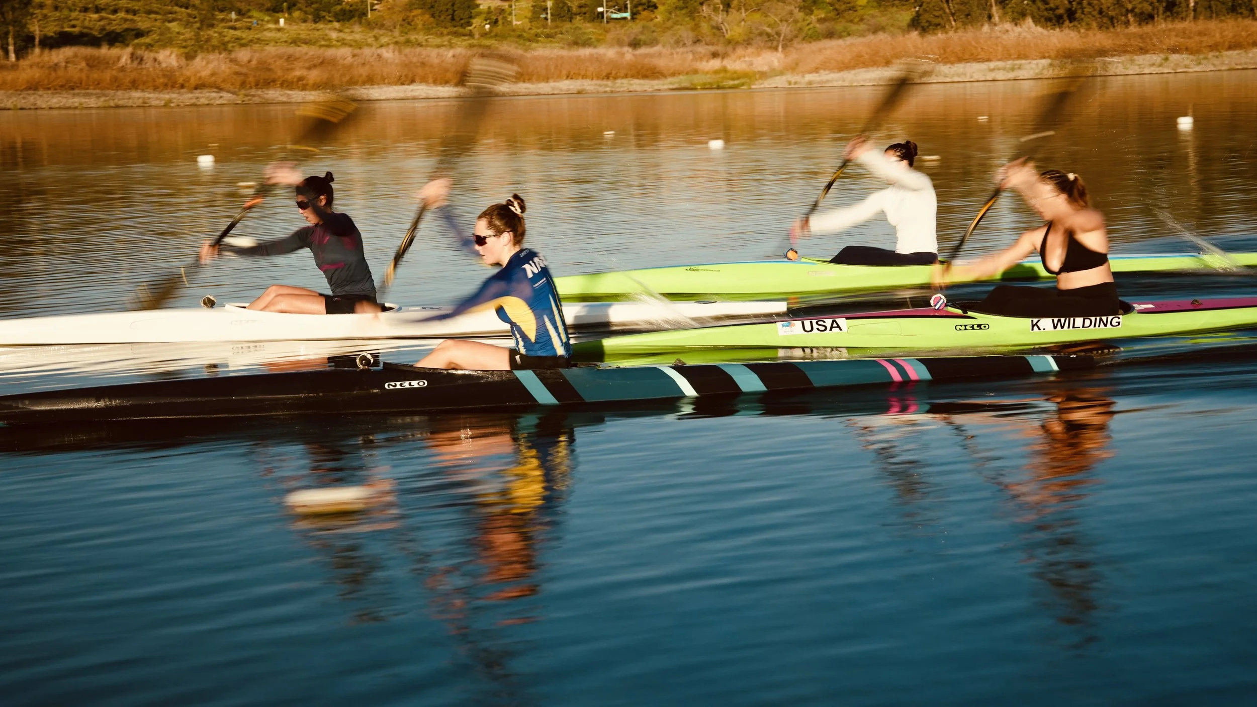 Four women in a racing kayak on a calm river during sunset, with two women in the front and two in the back, all wearing sunglasses and athletic clothing, paddling vigorously.