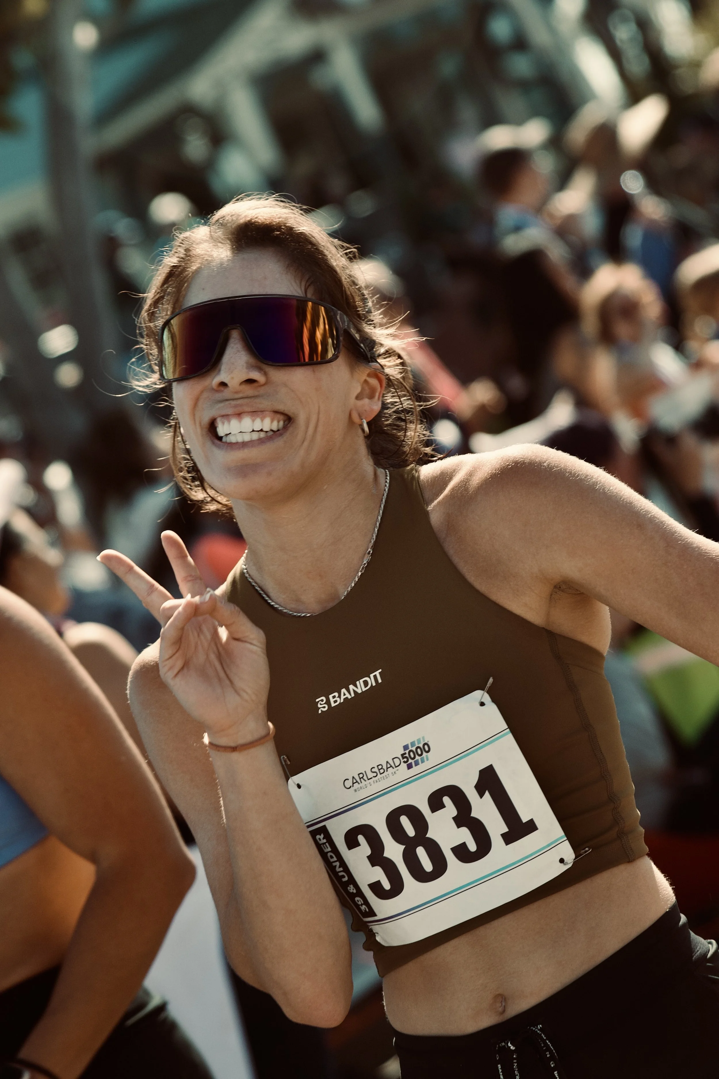 Smiling female runner wearing sunglasses, a brown sports top, and a race bib, in a crowded outdoor race event.