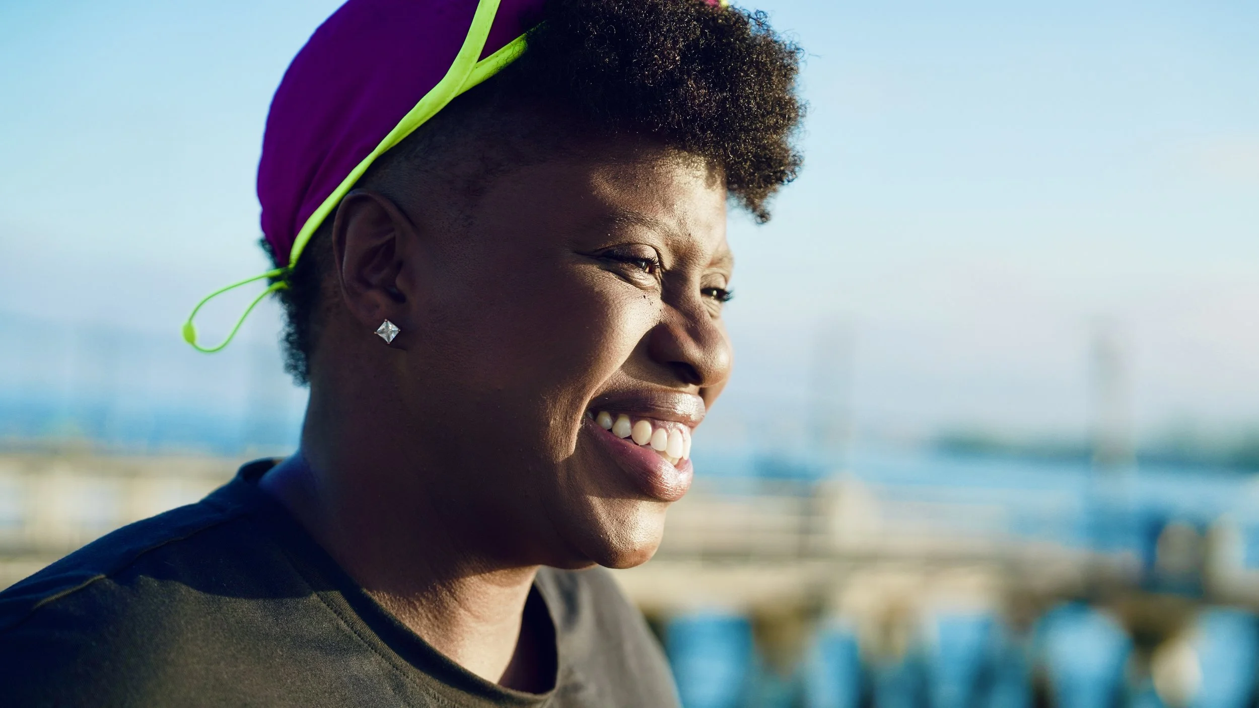 Close-up of an African American woman with a bright smile, short curly hair, wearing diamond earrings and a pink headwrap, outdoors with a blurred background of a beach and ocean.