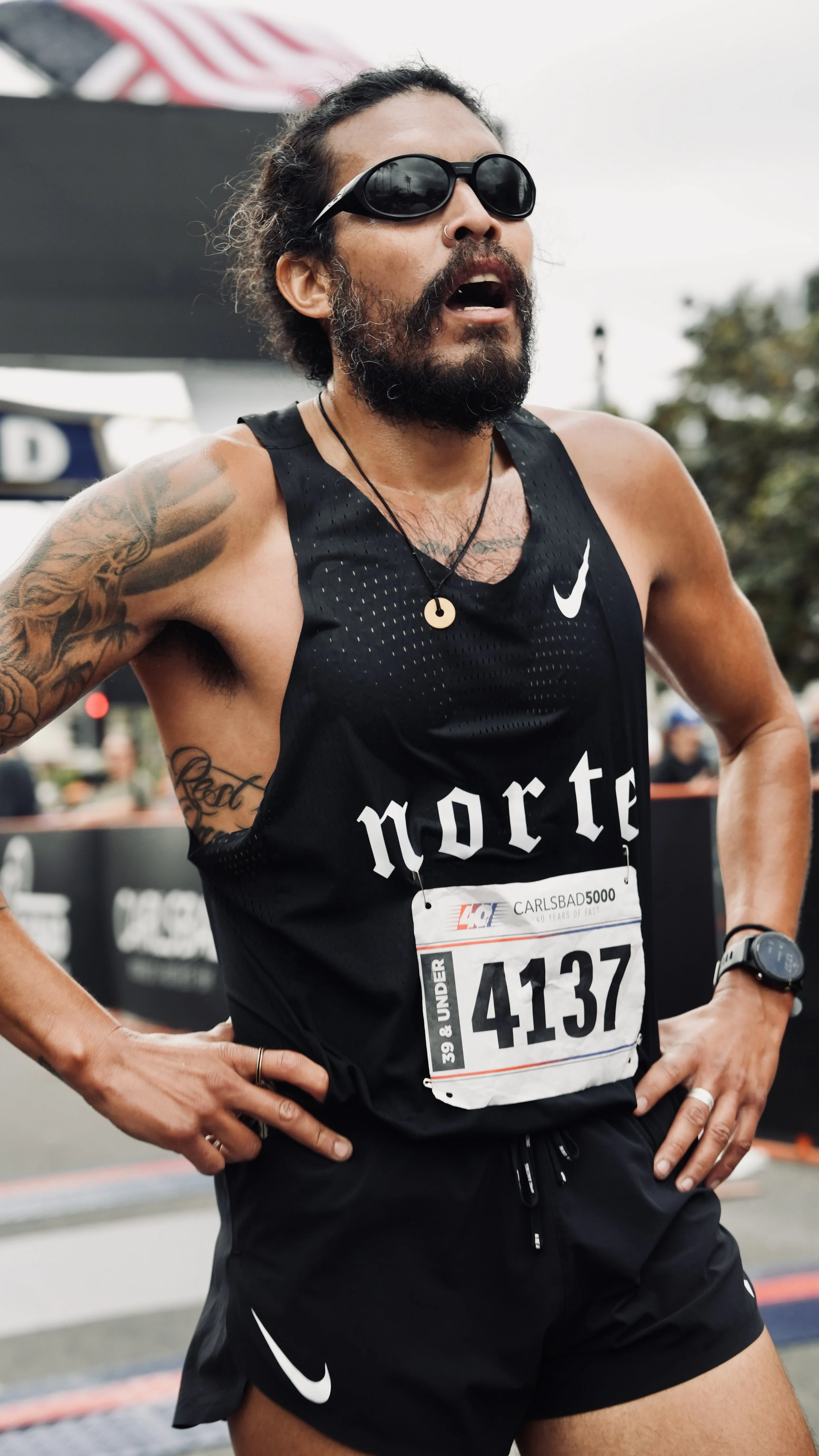 A male marathon runner with sunglasses and tattoos on his arm, wearing a black Nike tank top and black shorts, mid-race during a marathon event.