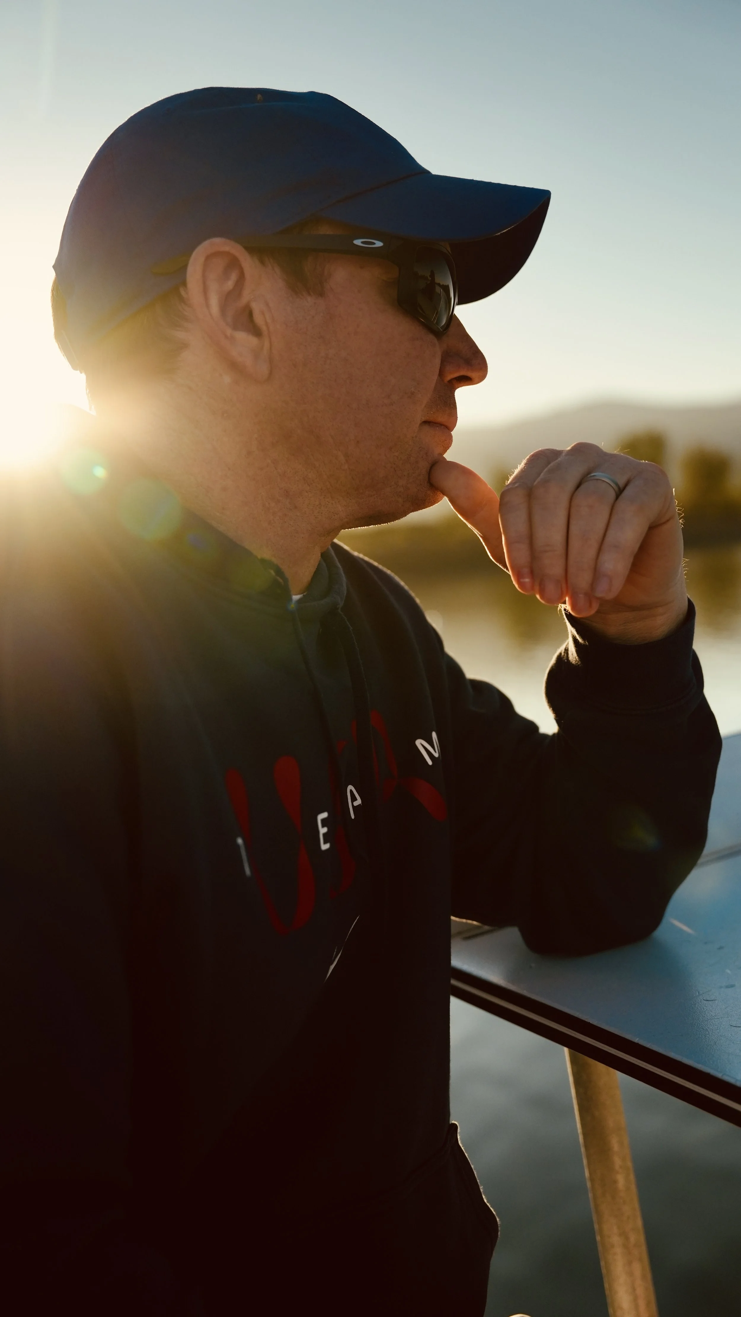 A man wearing a blue cap and sunglasses is standing outdoors during sunset, with one hand raised to his face, near his mouth, in a thoughtful pose, near a railing, with a body of water and mountains in the background.