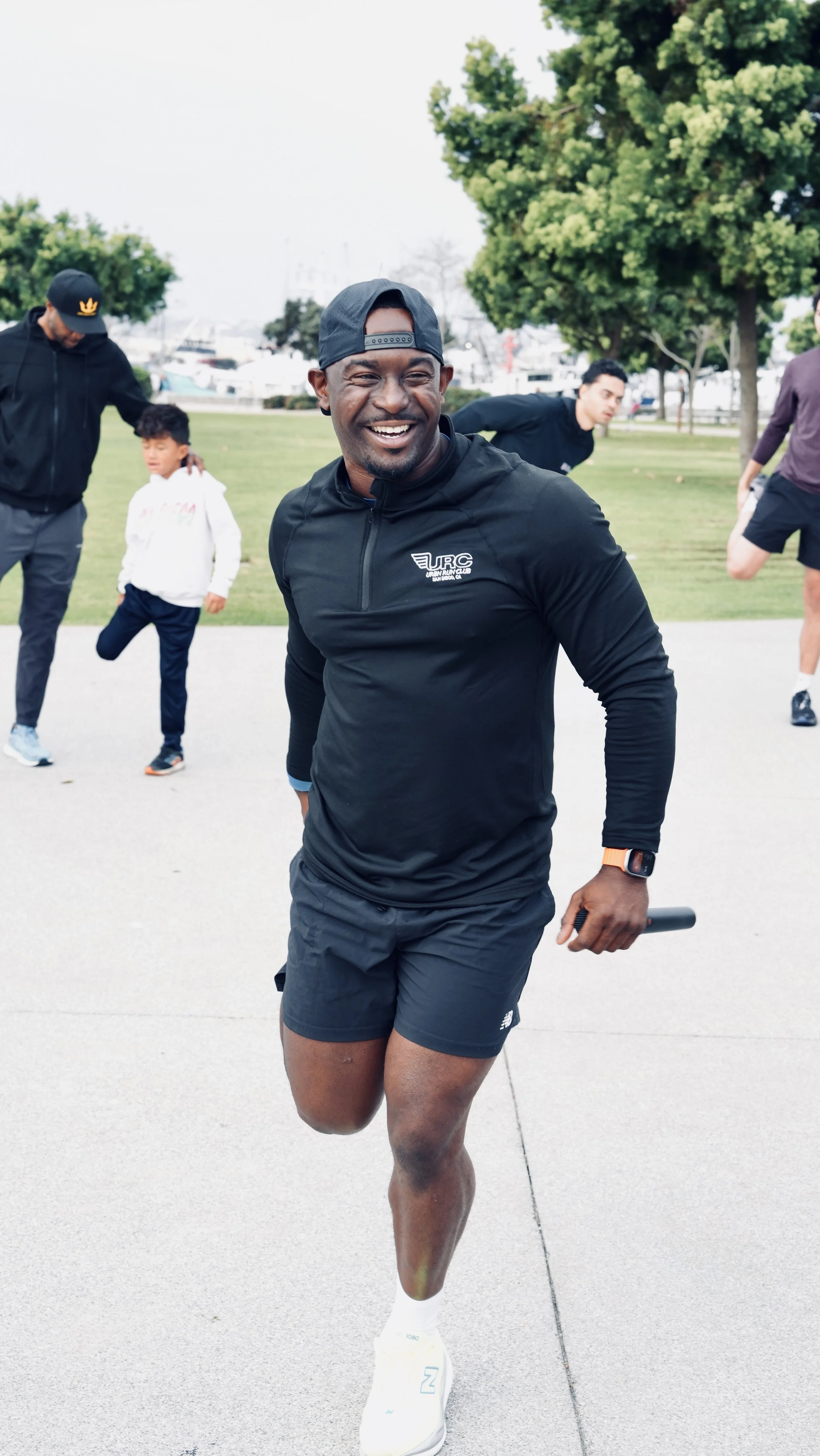 A man smiling while running outdoors in a park with green trees and a marina in the background.