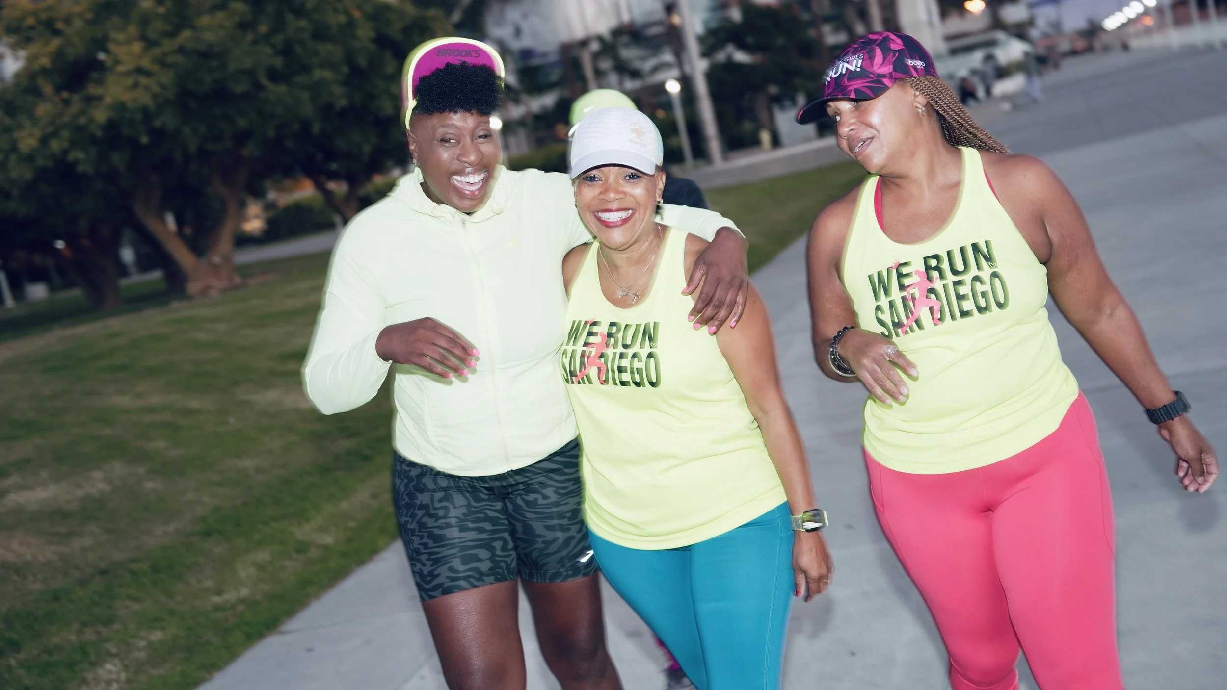 Three women joyfully running together in a park, wearing bright athletic clothing and smiling.