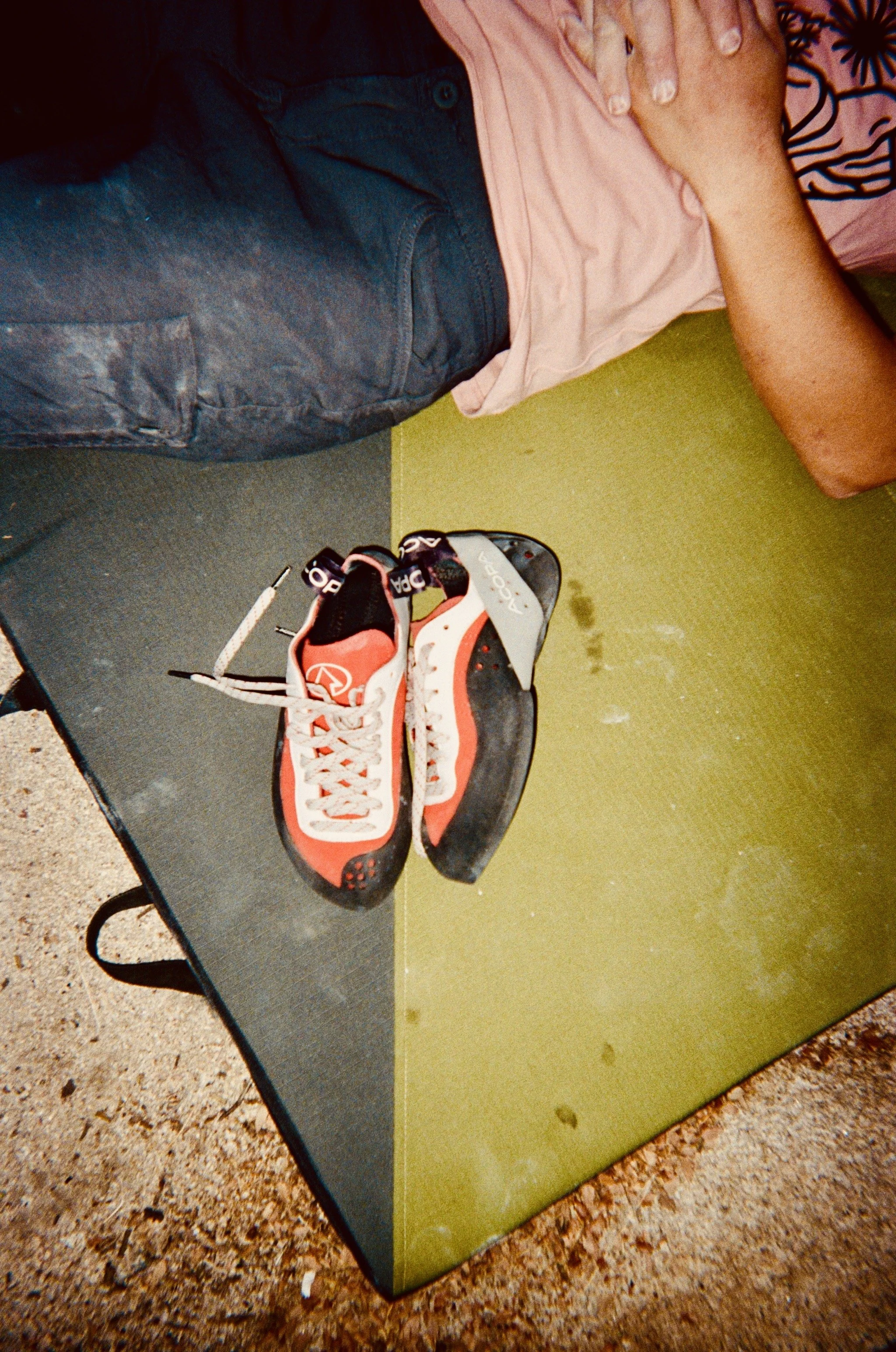 Climbing shoes on a green and black gym mat, with a person's arm resting on a pink shirt and a dark glove or piece of clothing partly visible.