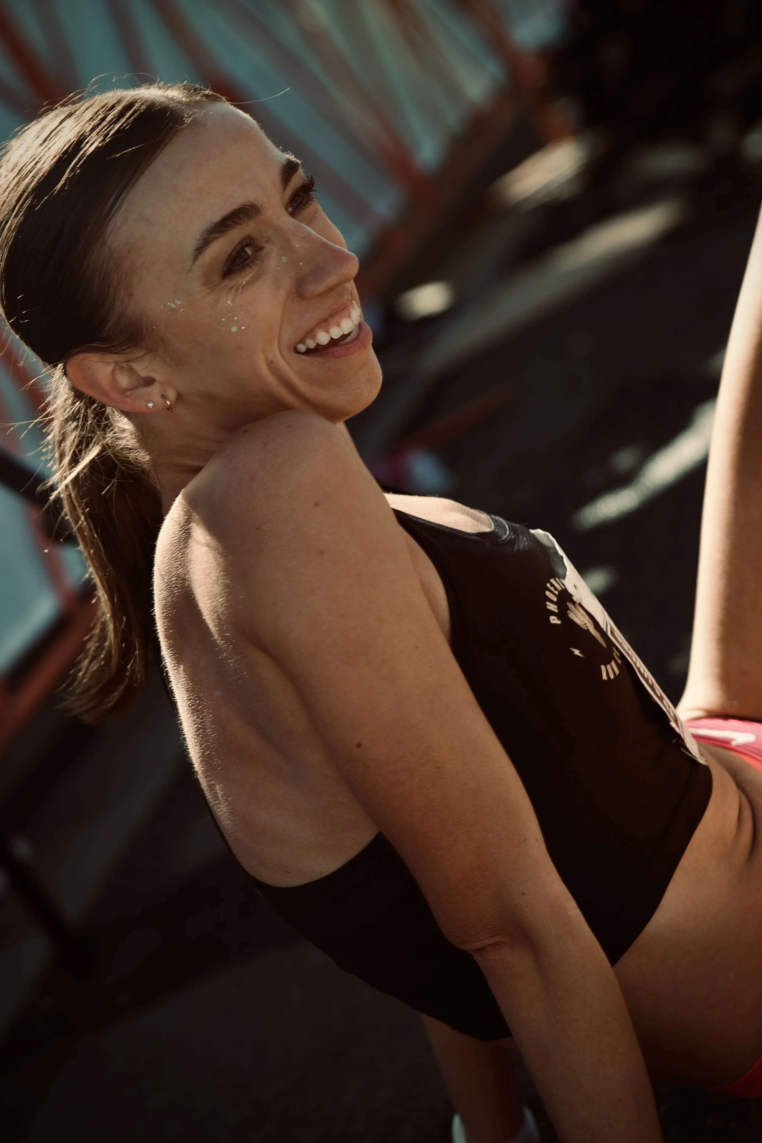 A woman with brown hair smiling and looking to the side, wearing a black sports top, in a setting with colorful blurred background.