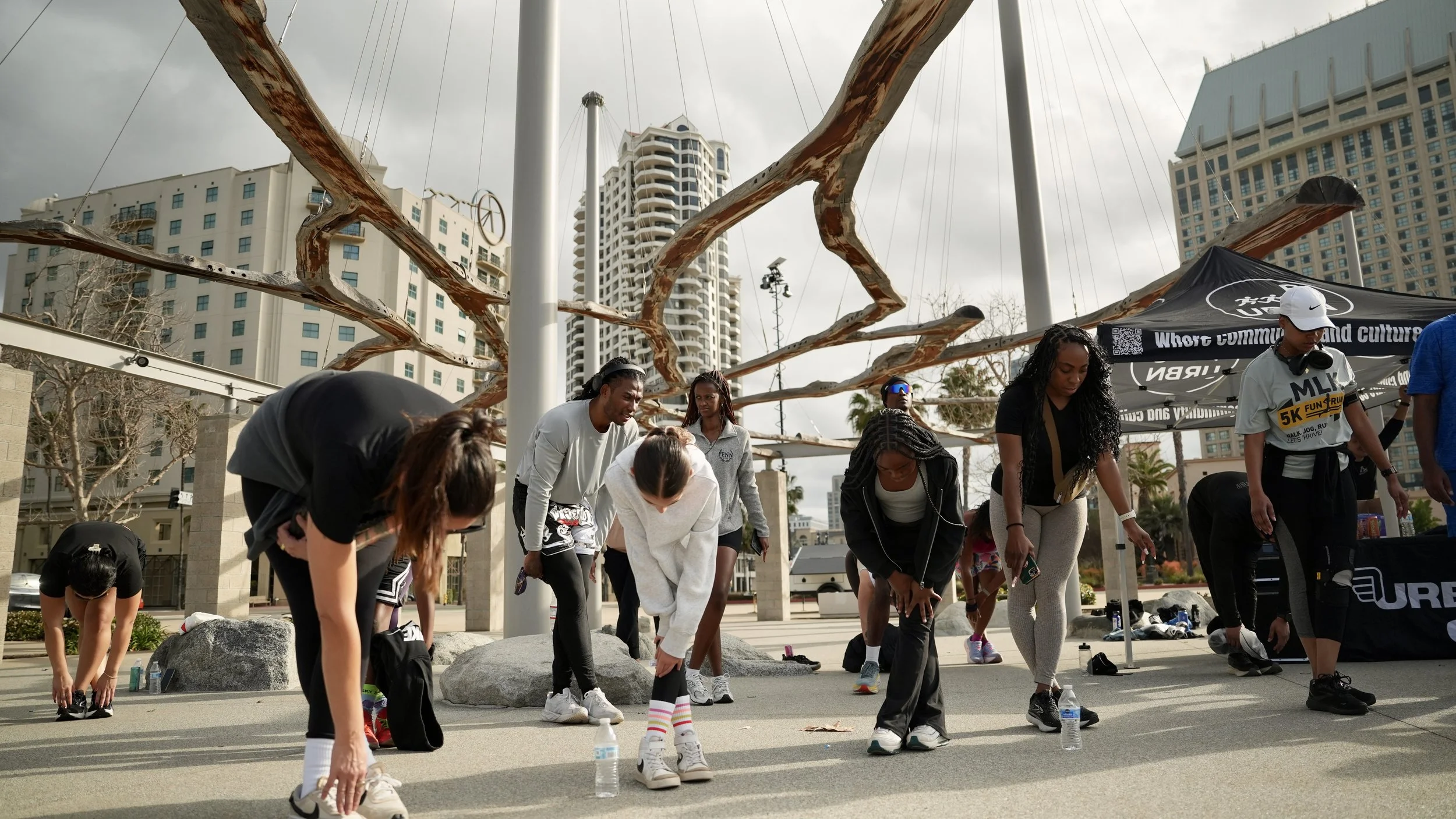 Group of people participating in an outdoor event, some bending down and others standing, with a large sculpture behind them and tall buildings in the background.