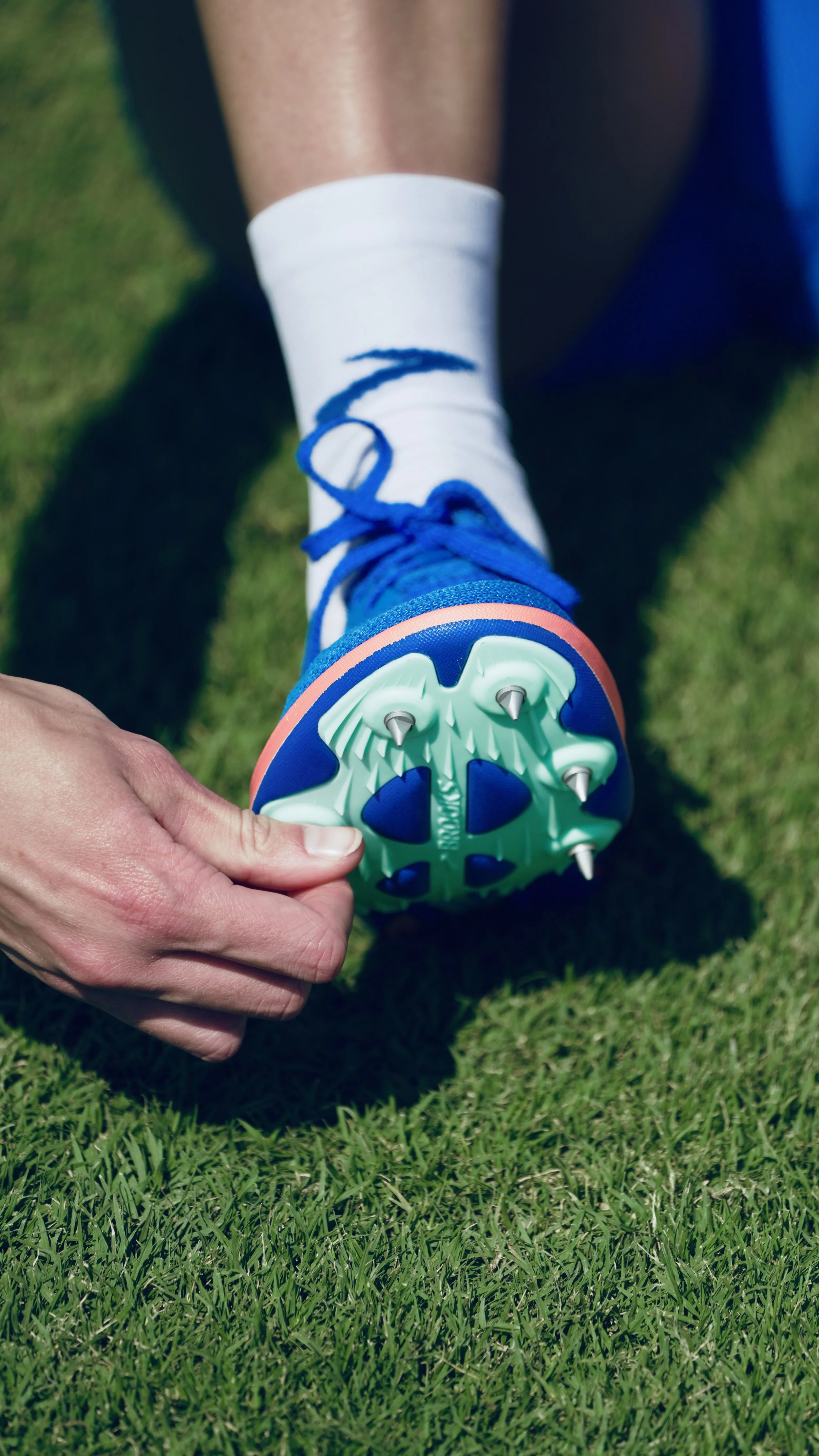 Close-up of a person's foot wearing a white sock with blue accents and a soccer cleat, on green grass field.