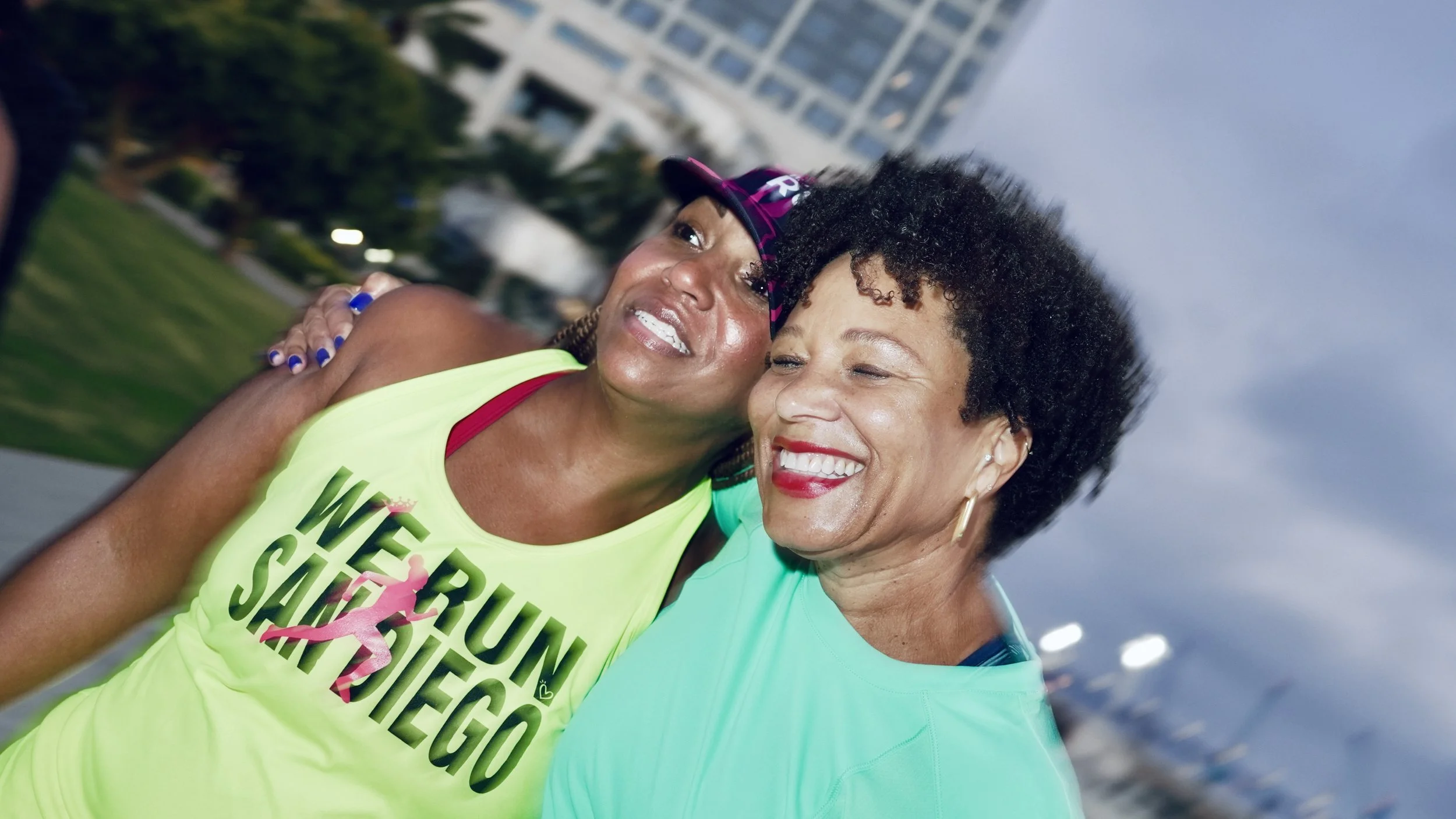 Two women hugging and smiling at a running event, with one wearing a neon yellow shirt that says "WE RUN SURVIVORS" and a pink ribbon graphic, and the other in a teal shirt, outdoors with buildings in the background.