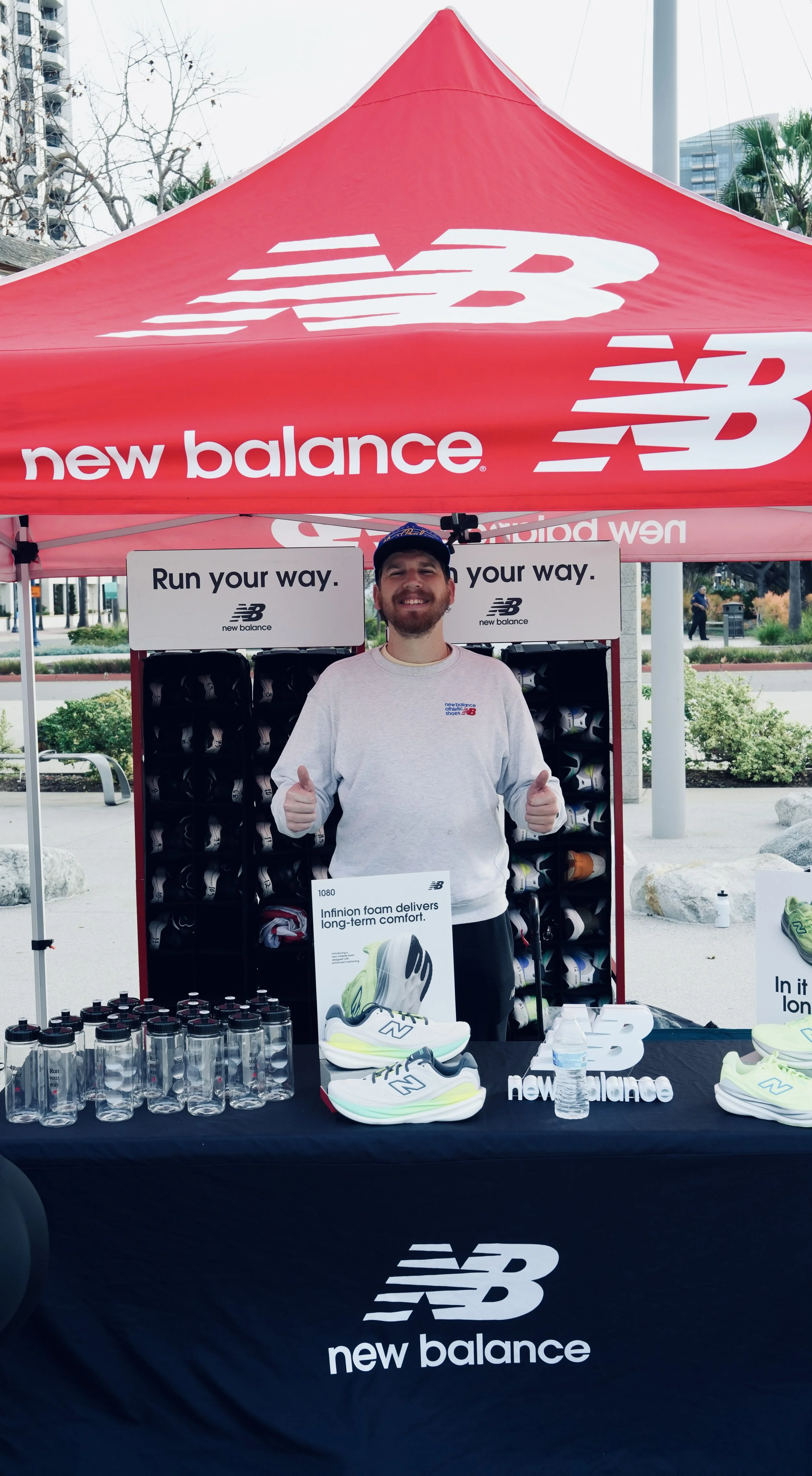A man standing under a red New Balance tent at an outdoor promotional event. The man is smiling and giving two thumbs up. The table in front of him displays New Balance sneakers, water bottles, and promotional materials. There are signs that say 'Run