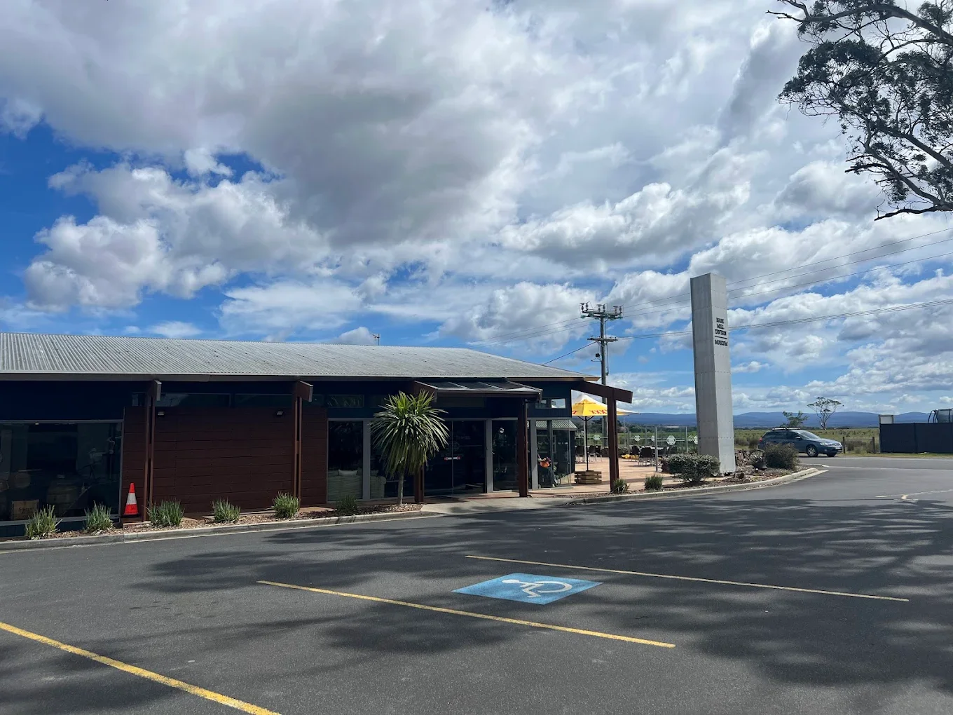 A modern building with a parking lot in front, including a designated handicapped parking space, under a partly cloudy sky with blue patches. There are some trees and bushes around the building and a tall sign structure nearby.