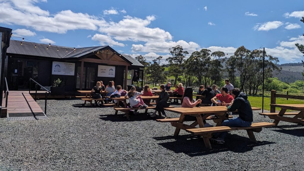 People sitting at picnic tables outside a building on a sunny day, surrounded by trees and hills.