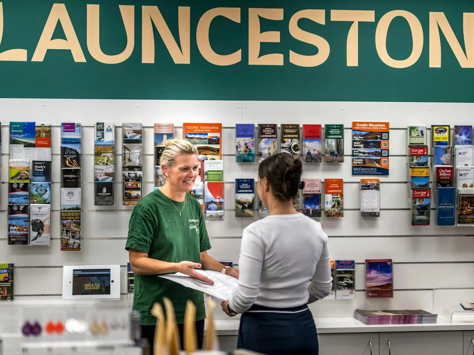 Two women talking in front of a wall with brochures and tourist information in Launceston, Tasmania.