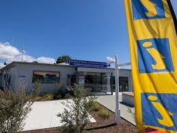 Exterior of a store with a blue sign, surrounded by plants and a yellow and blue banner.